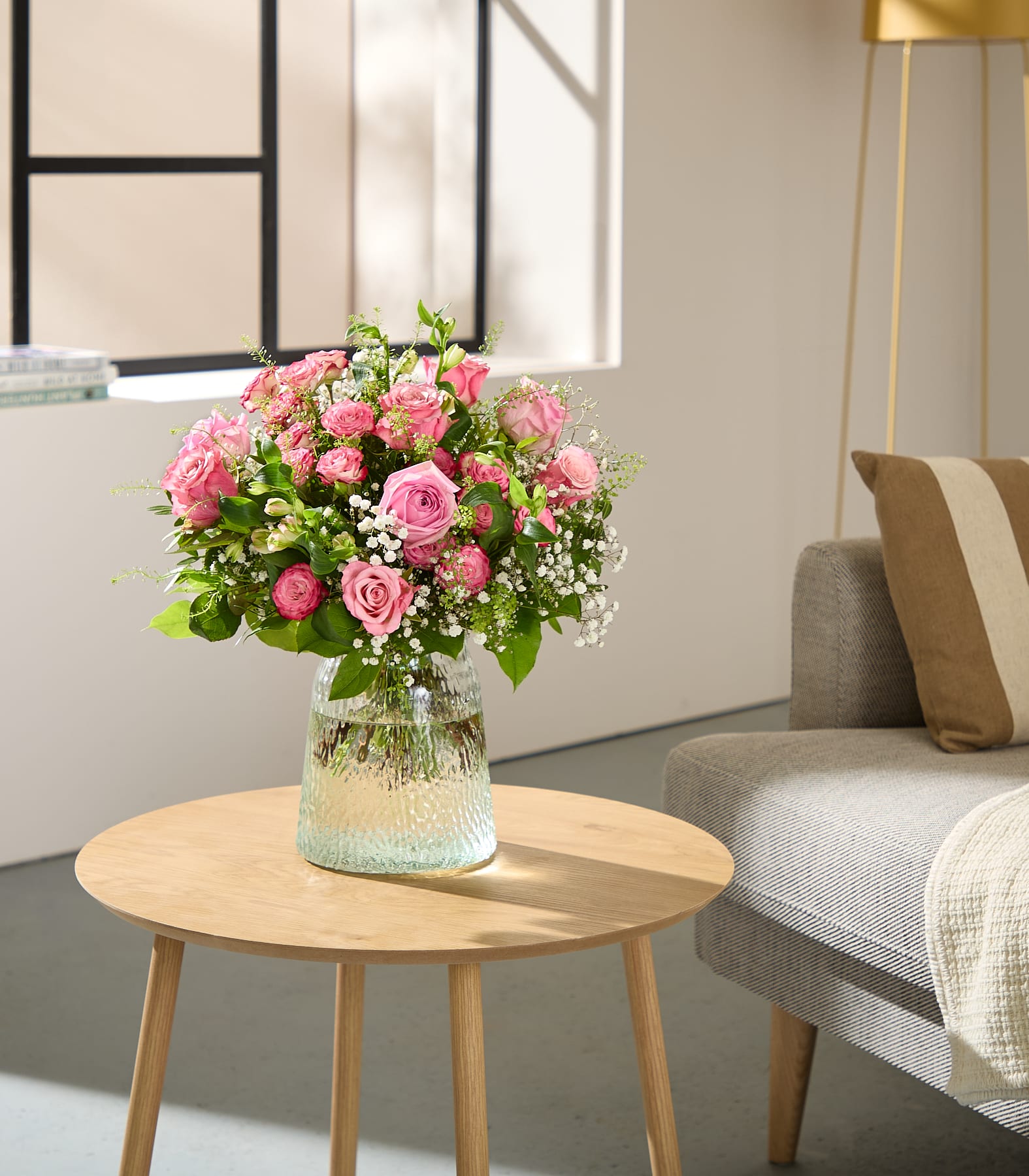 Eine junge Frau mit langen braunen Haaren hält vor einem schlichten weißen Hintergrund einen leuchtenden Strauß aus rosa Rosen und anderen Blumen.