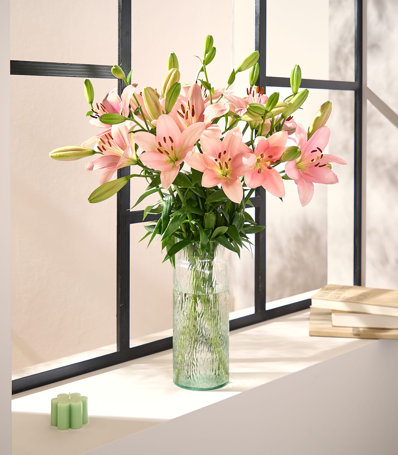 Auf einem Fensterbrett steht eine Vase mit leuchtend pinkfarbenen Lilien. Im Hintergrund sind Bücher und ein kleiner grüner Gegenstand zu sehen, vor dem Hintergrund einer verschneiten Außenszene, die man durch das Fenster erblickt.