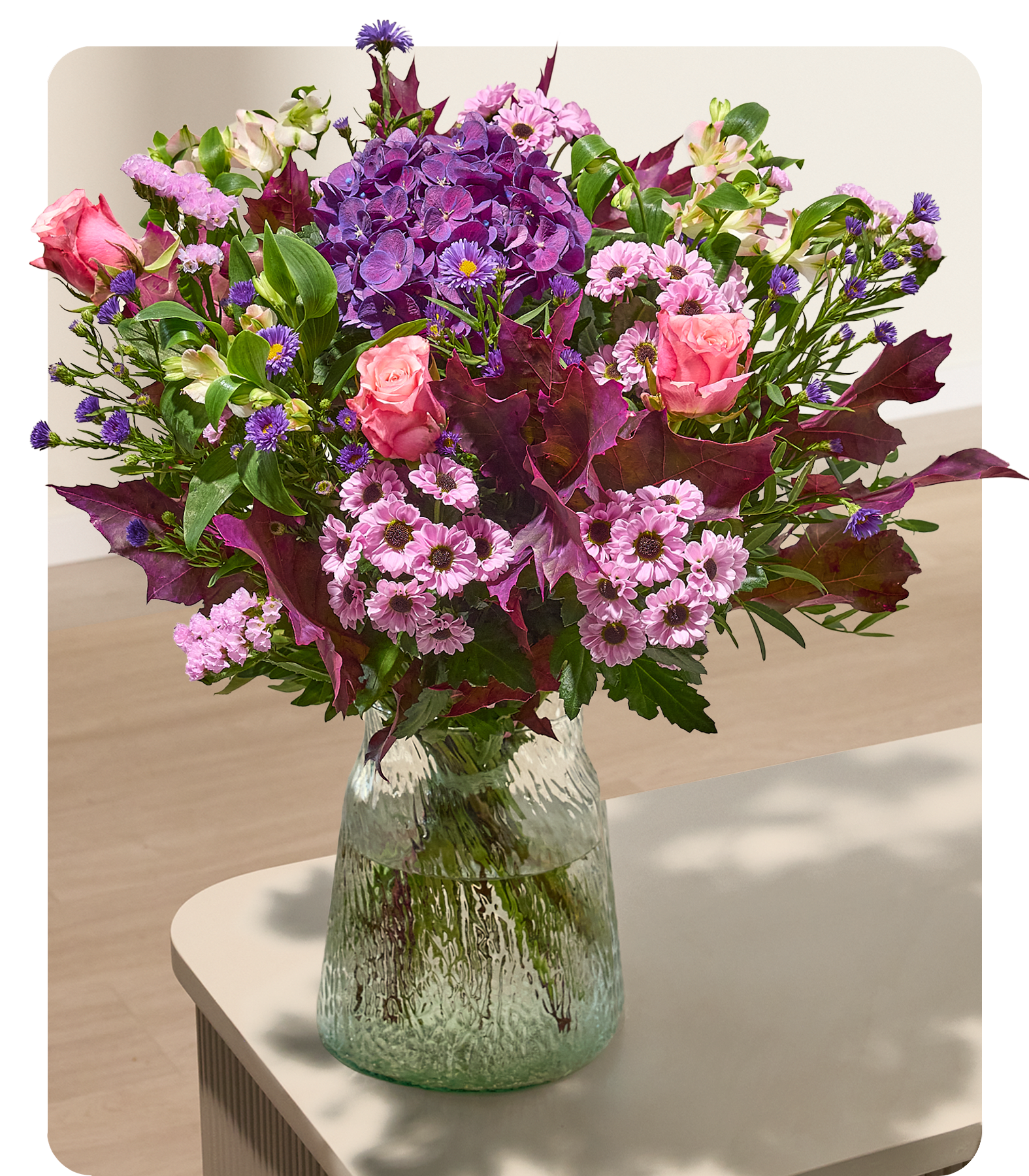 Ein farbenfroher Strauß aus rosa und violetten Blumen, darunter Rosen und Hortensien, ist in einer strukturierten Glasvase auf einem hellen Tisch arrangiert; im Hintergrund sind ein verschwommener Holzboden und eine Holzwand zu sehen. (Beschreibungstext KI-generiert)