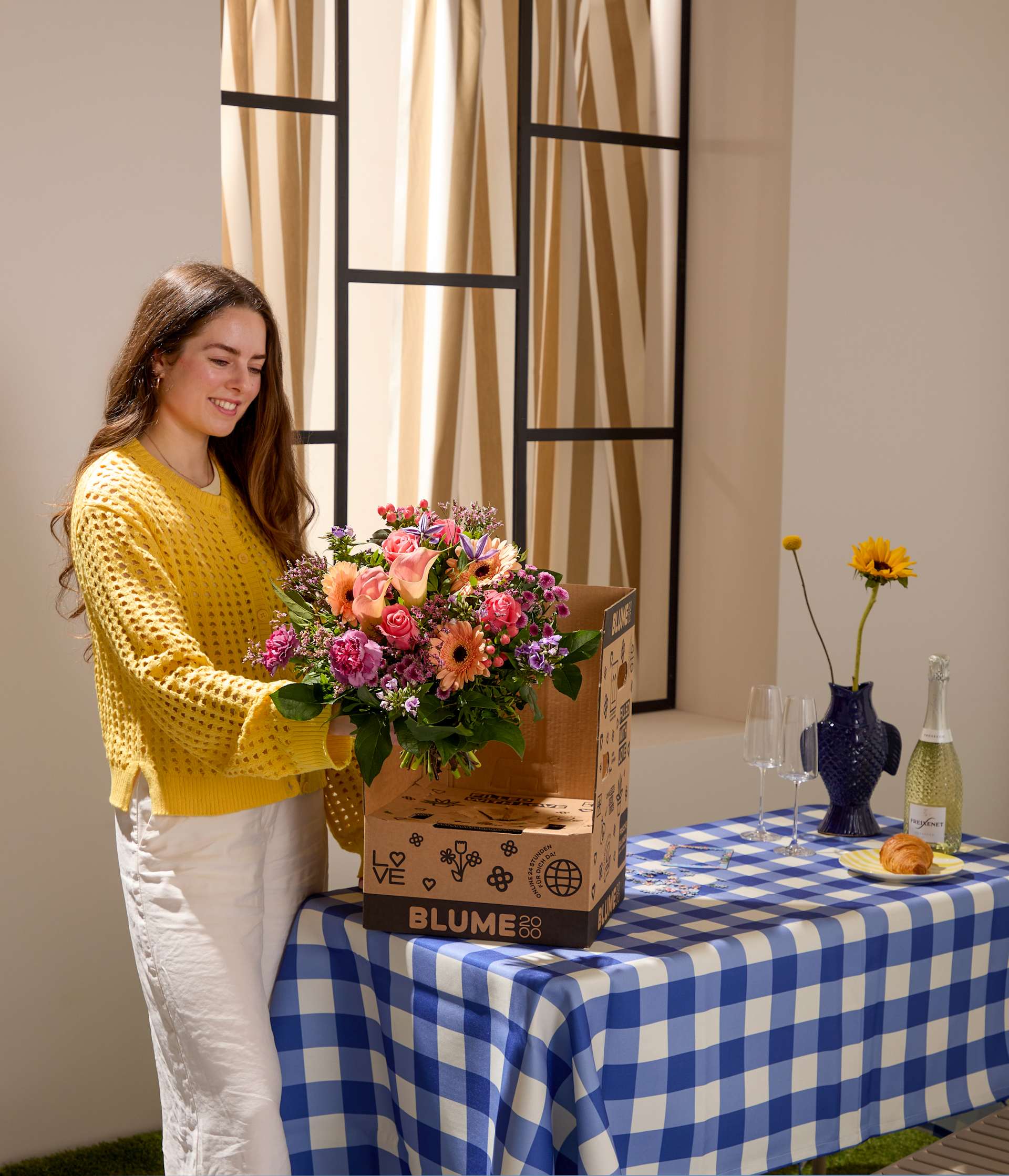 Eine junge Frau in einem gelben Pullover hält einen Blumenstrauß neben einem Lieferkarton; im Hintergrund sind eine karierte Tischdecke, Weingläser und eine Flasche Champagner aufgestellt.