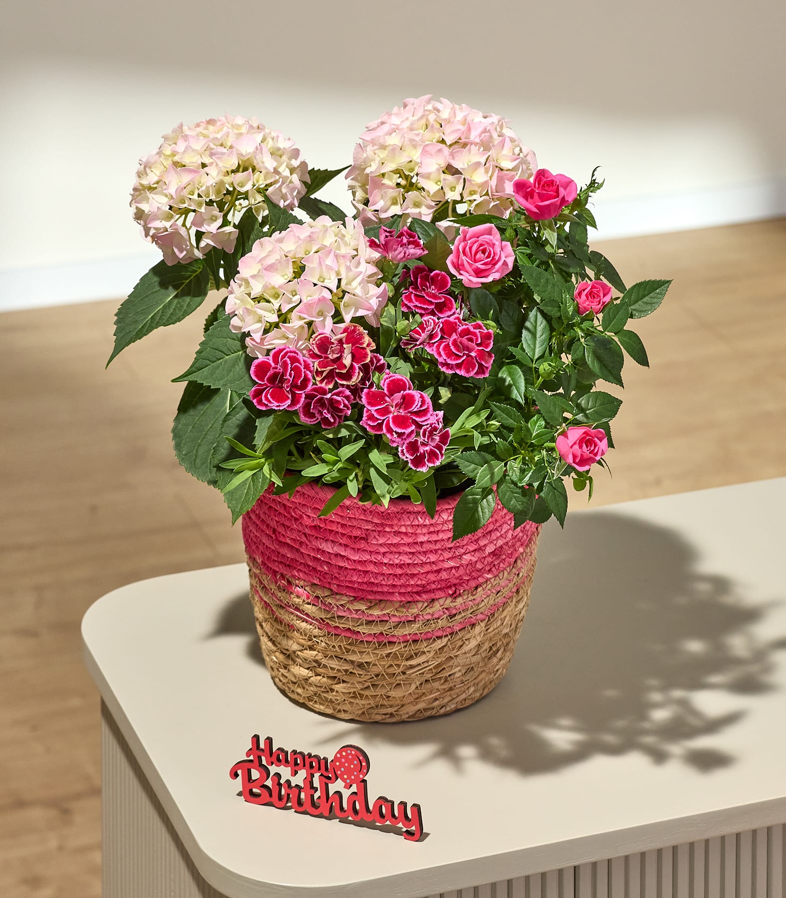 Ein rosafarbener, mit Stroh geflochtener Topf ist übervoll mit leuchtend pinkfarbenen Rosen und Hortensien, während ein rotes Schild mit der Aufschrift „Happy Birthday“ auf einem hellen Tisch vor einem verschwommenen Hintergrund steht.