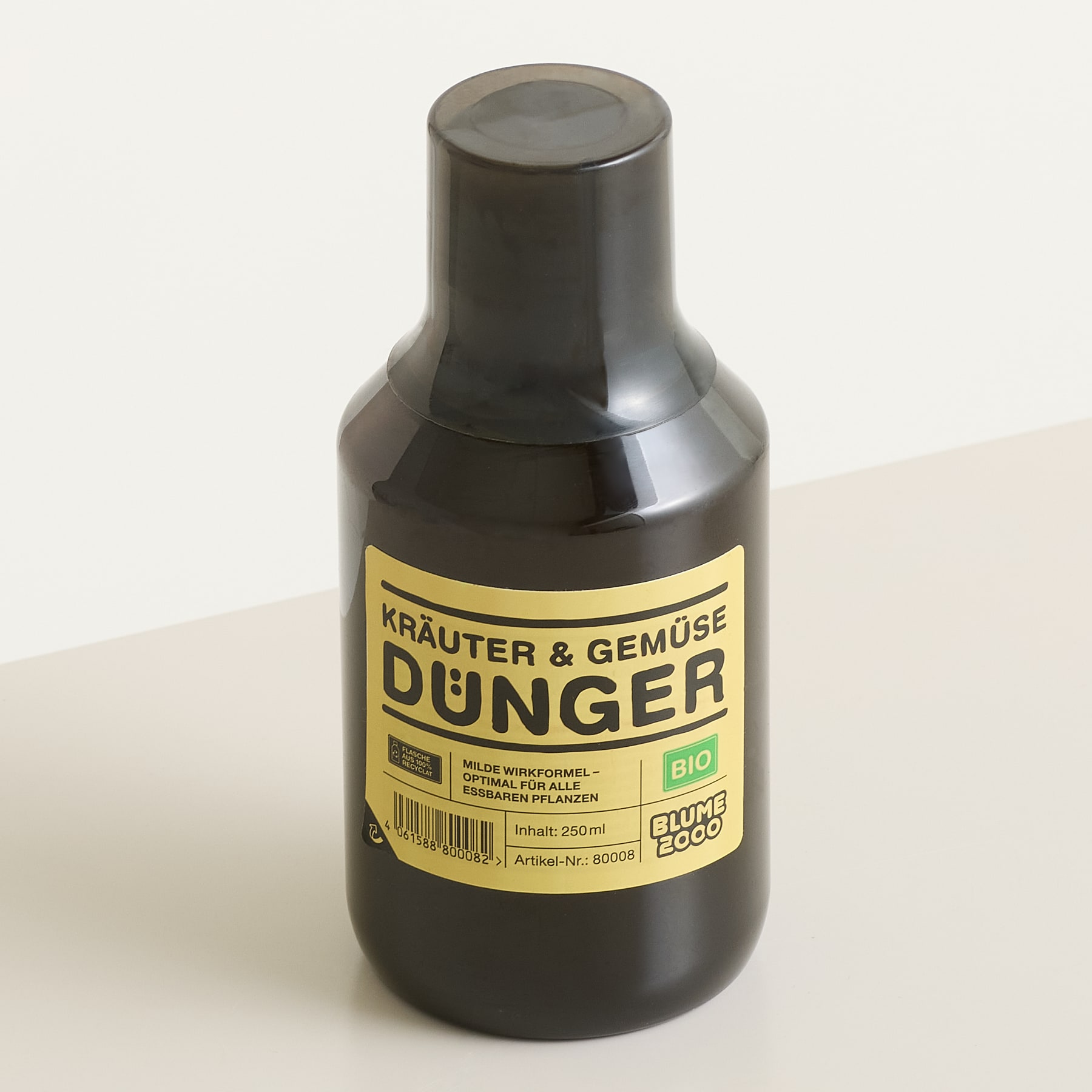 Im Vordergrund steht vor einem schlichten weißen Hintergrund eine dunkelbraune Flasche „Kräuter & Gemüse Dünger“ mit gelbem Etikett.