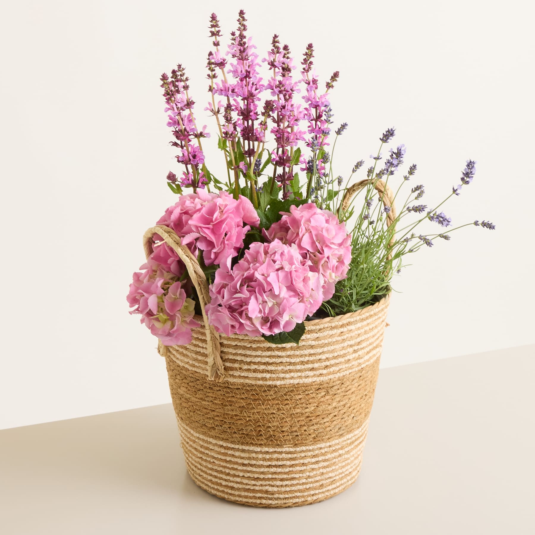 Ein geflochtener Korb, gefüllt mit rosa Hortensien und lila Blüten, steht auf einer neutralen Fläche vor einem schlichten Hintergrund.