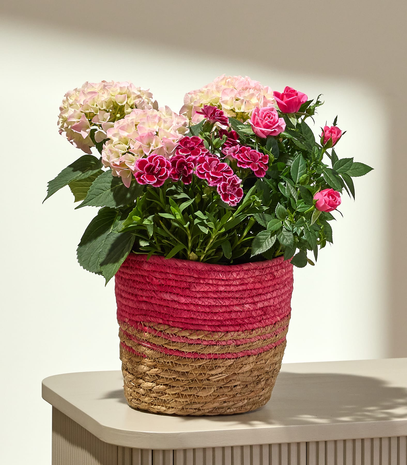 Ein farbenfroher Strauß aus rosa und roten Blumen, darunter Rosen und Dahlien, ist in einem geflochtenen Korb auf einer hellen Unterlage arrangiert.