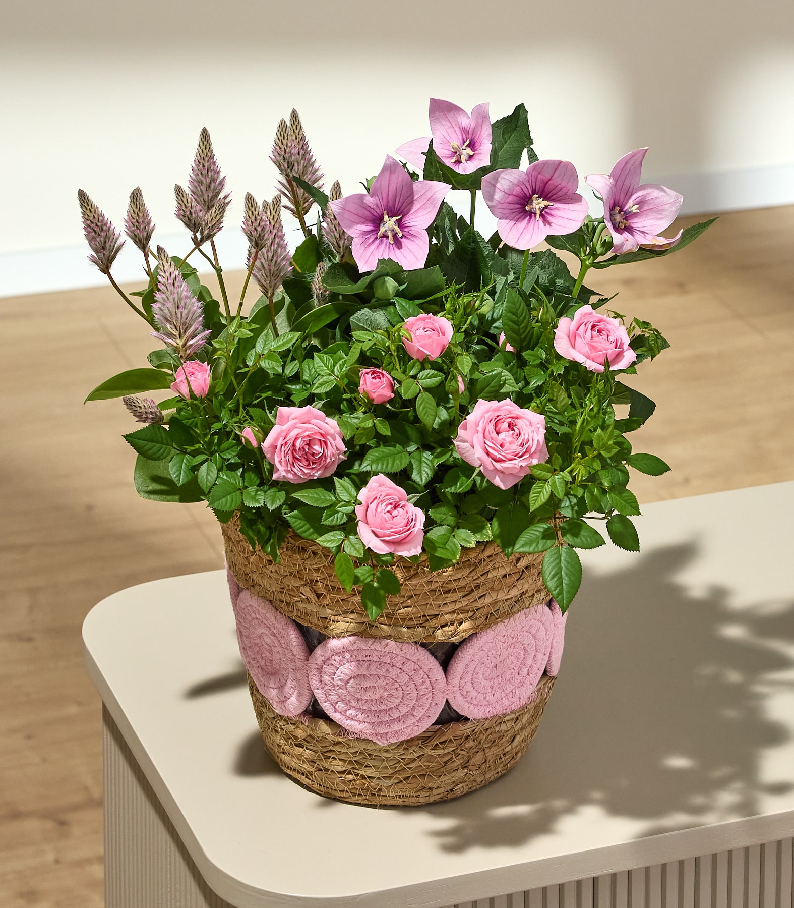 Das Bild zeigt ein lebhaftes Arrangement aus rosa Rosen, umgeben von üppigem grünem Laub und hohen, schlanken rosa und braunen Blütenstielen, vor einem schlichten, hellen Hintergrund.