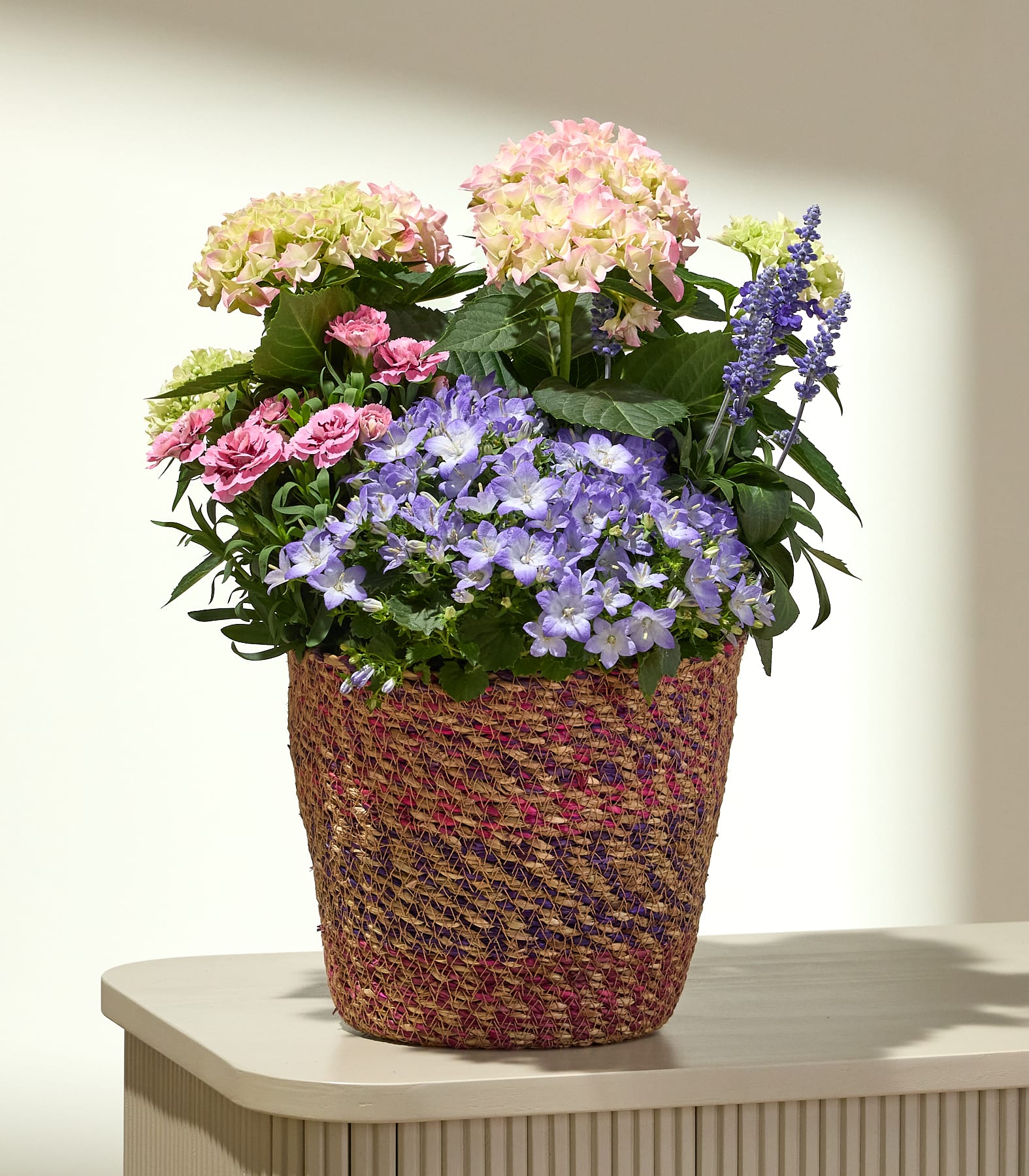 Ein geflochtener Korb ist übervoll mit einem lebhaften Arrangement aus rosa Rosen, lila und blauen Glockenblumen sowie rosa und gelben Hortensien vor einem sanft beleuchteten, neutralen Hintergrund.