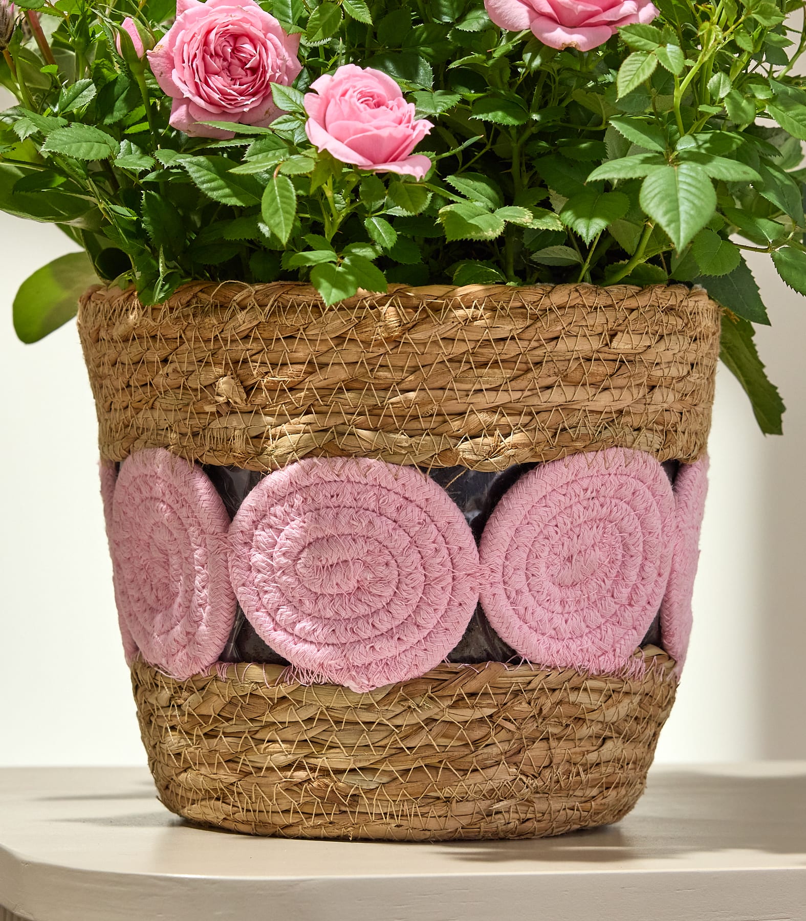Ein geflochtener Korb mit grünen Blättern und rosa, flauschigen Gegenständen darin steht auf einer hellen Oberfläche.