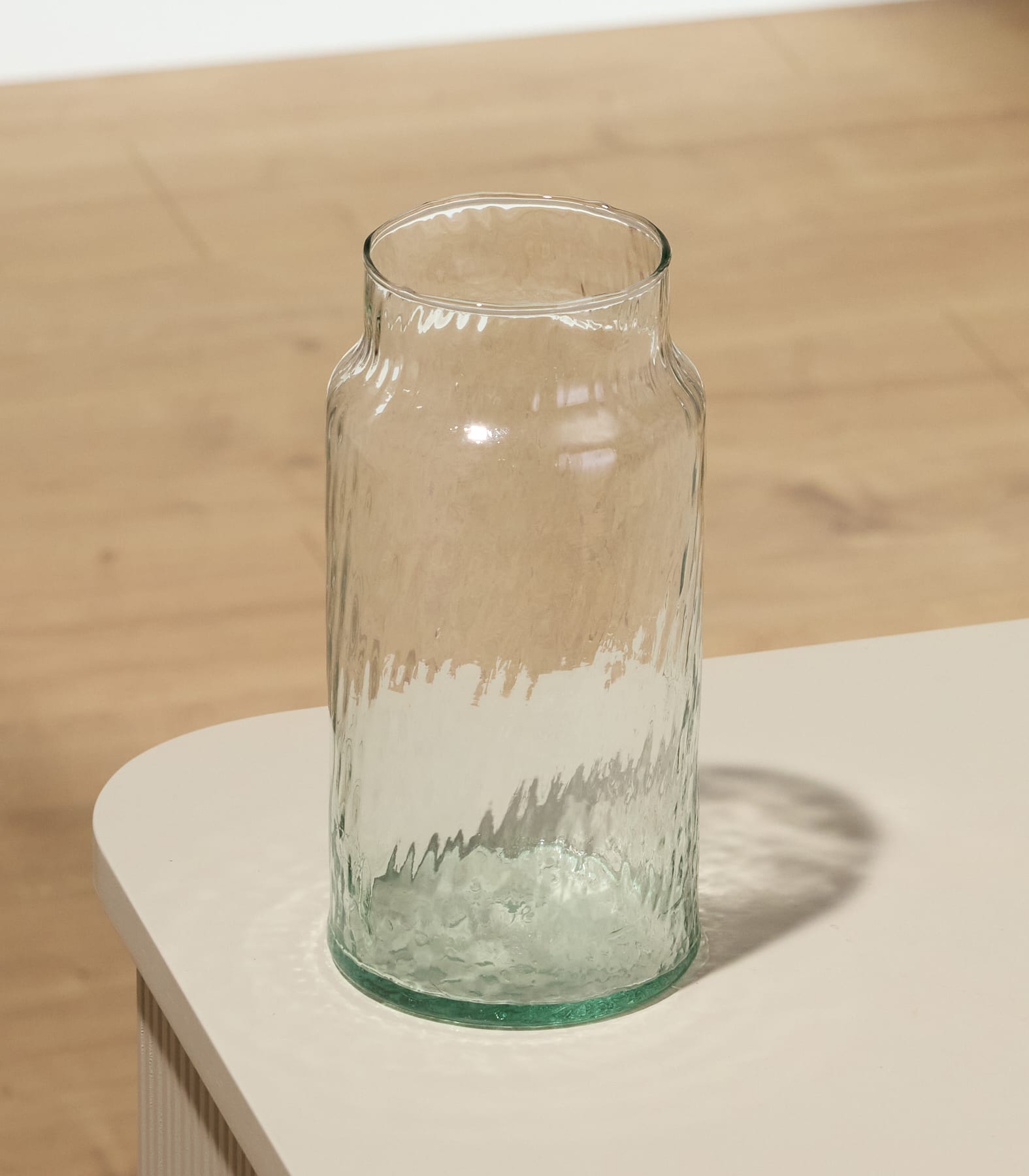 Eine durchsichtige Glasvase oder ein Krug mit zylindrischer Form und leicht nach oben verjüngter Öffnung steht auf einer hellen Oberfläche vor einem schlichten weißen Hintergrund.