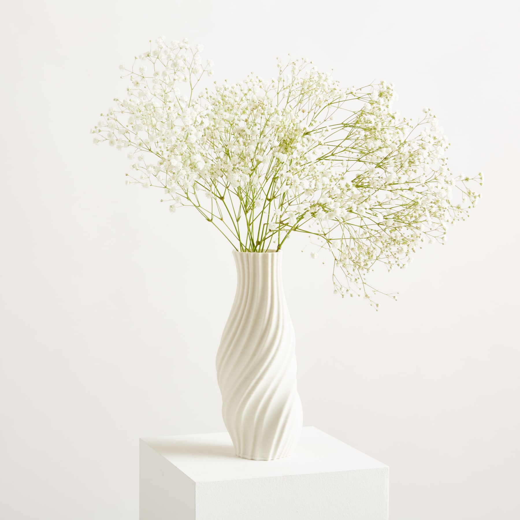 Eine weiße Vase mit zarten weißen Blüten steht auf einem weißen Sockel vor einem schlichten weißen Hintergrund.