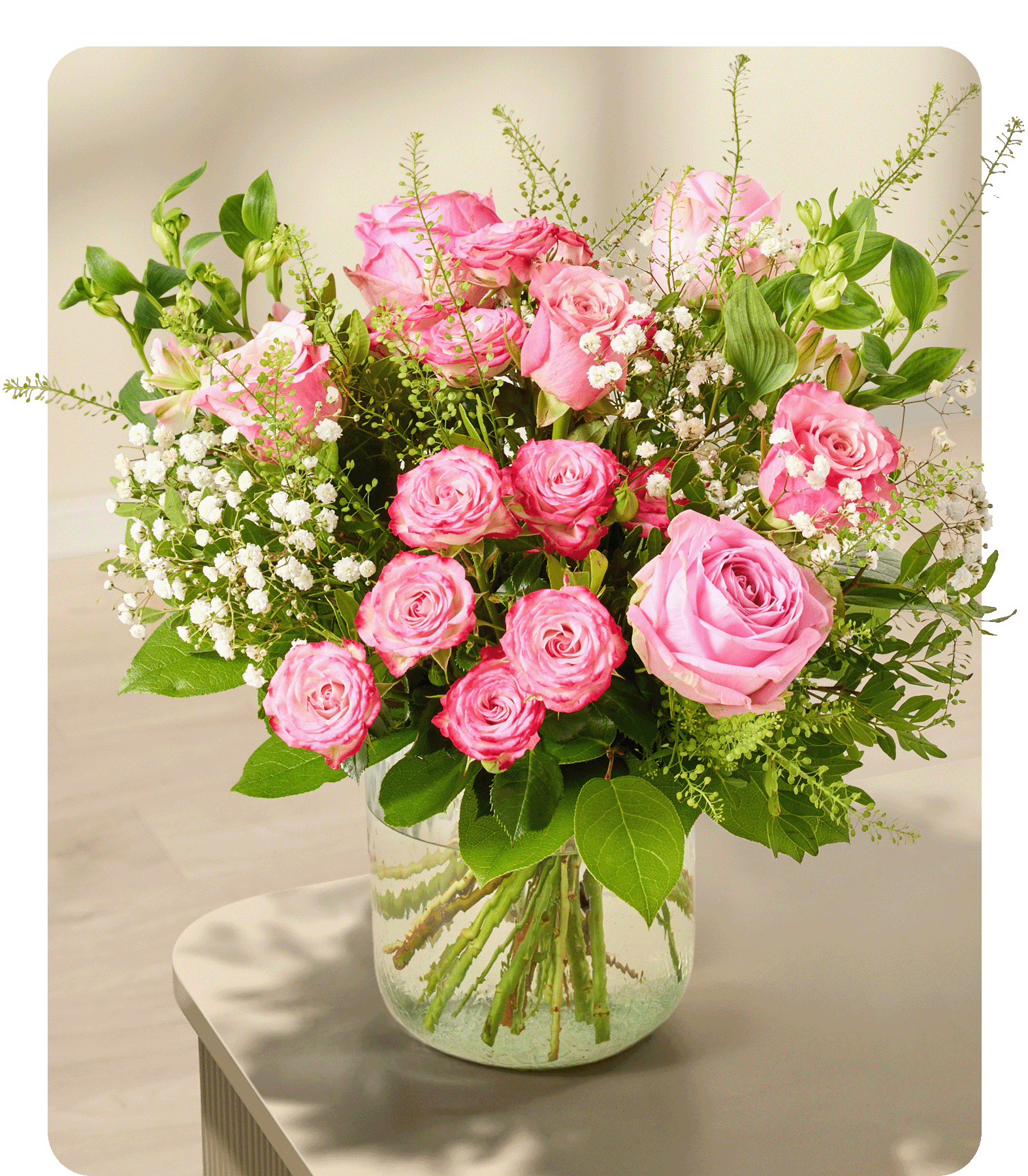 Ein Strauß rosa Rosen und Schleierkraut füllt eine durchsichtige Glasvase auf hellem Untergrund vor einem sanft verschwommenen Hintergrund.