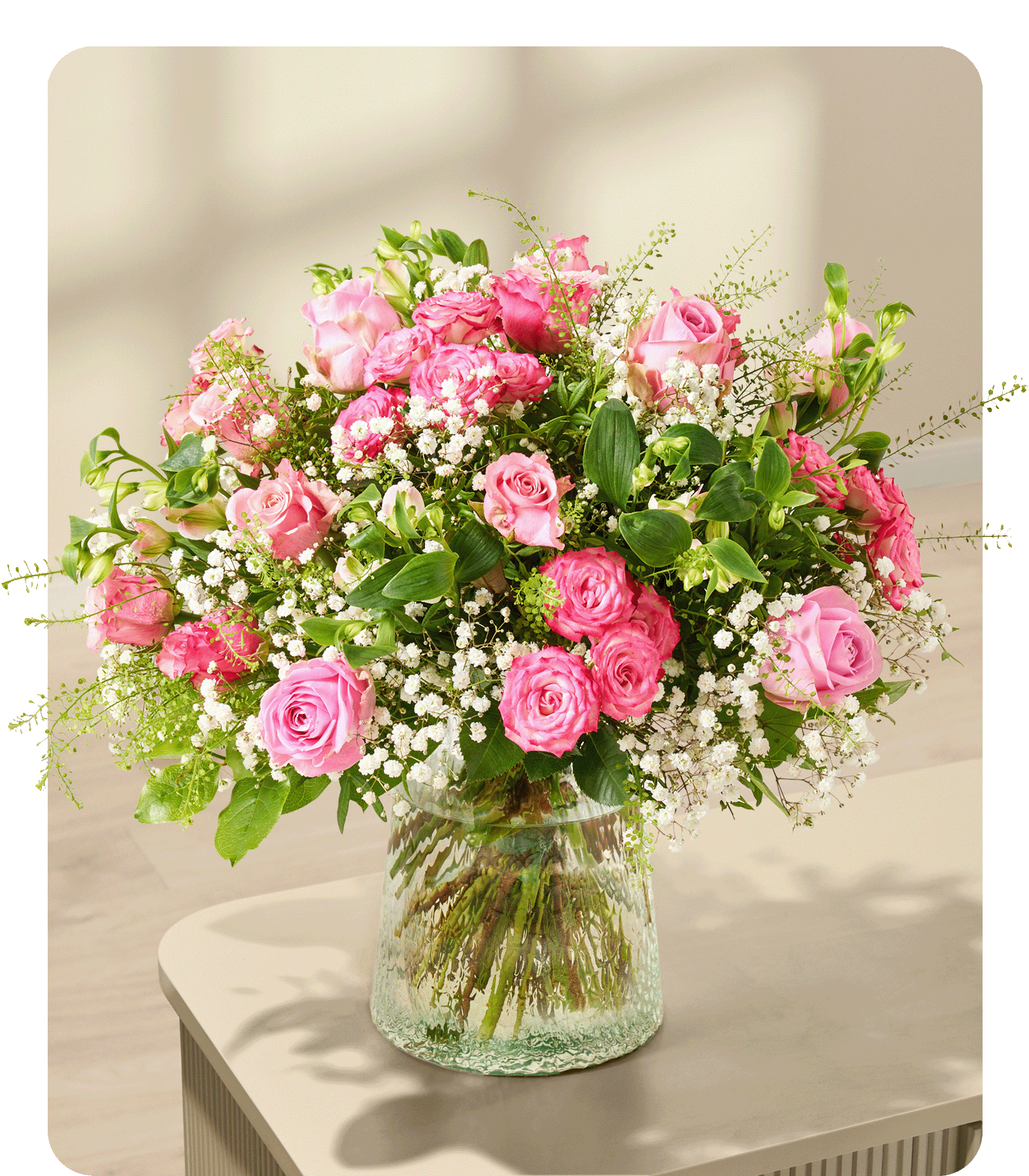 Ein üppiger Strauß aus rosa Rosen und Schleierkraut füllt eine strukturierte Glasvase auf hellem Untergrund vor einem sanft verschwommenen Hintergrund aus Licht- und Schattenspielen.