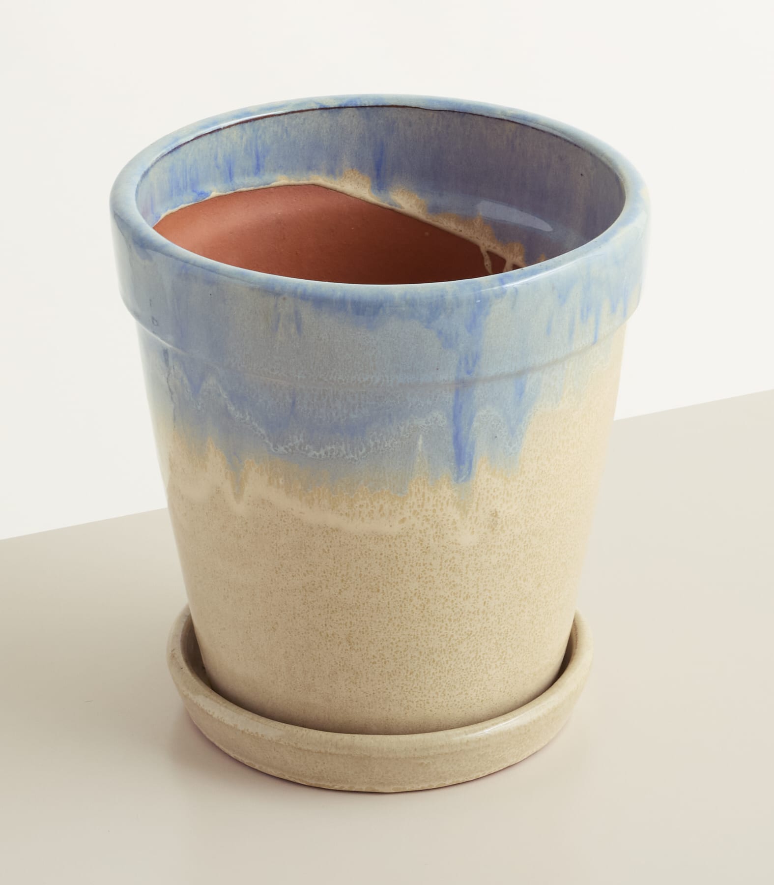 Ein hellblauer und beiger Keramiktopf mit passender Untertasse steht auf einer hellen Oberfläche vor einem sanften, cremeweißen Hintergrund.