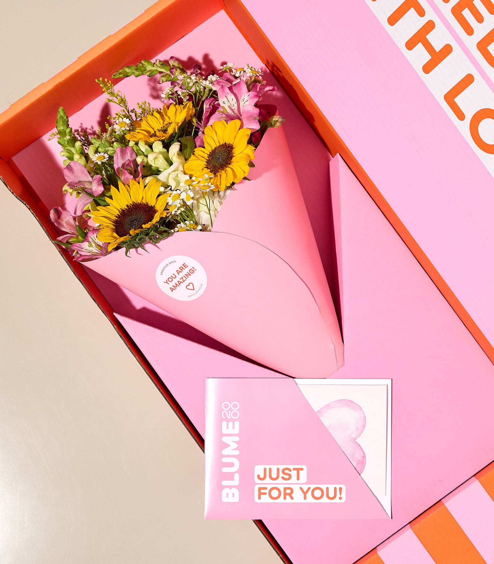 Ein Strauß Sonnenblumen und anderer Blumen wird in einem rosafarbenen Kegel innerhalb einer leuchtend orangefarbenen Schachtel präsentiert, begleitet von einer Karte mit der Aufschrift „Nur für dich!“.