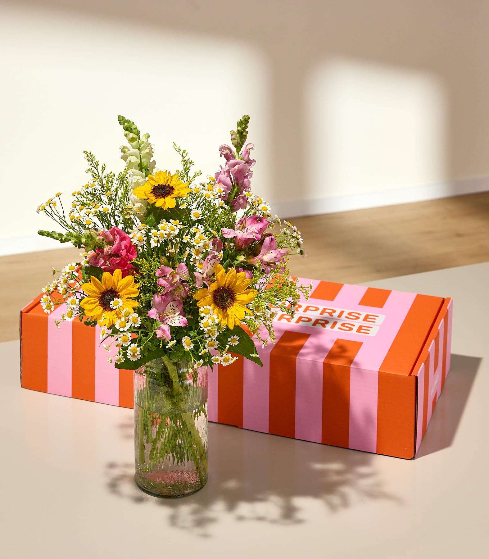 Ein leuchtender Strauß Sonnenblumen und Wildblumen steht in einer Glasvase vor einer rosa-orange gestreiften Geschenkbox; im Hintergrund fallen sanfte Schatten auf eine helle Oberfläche und eine Wand.