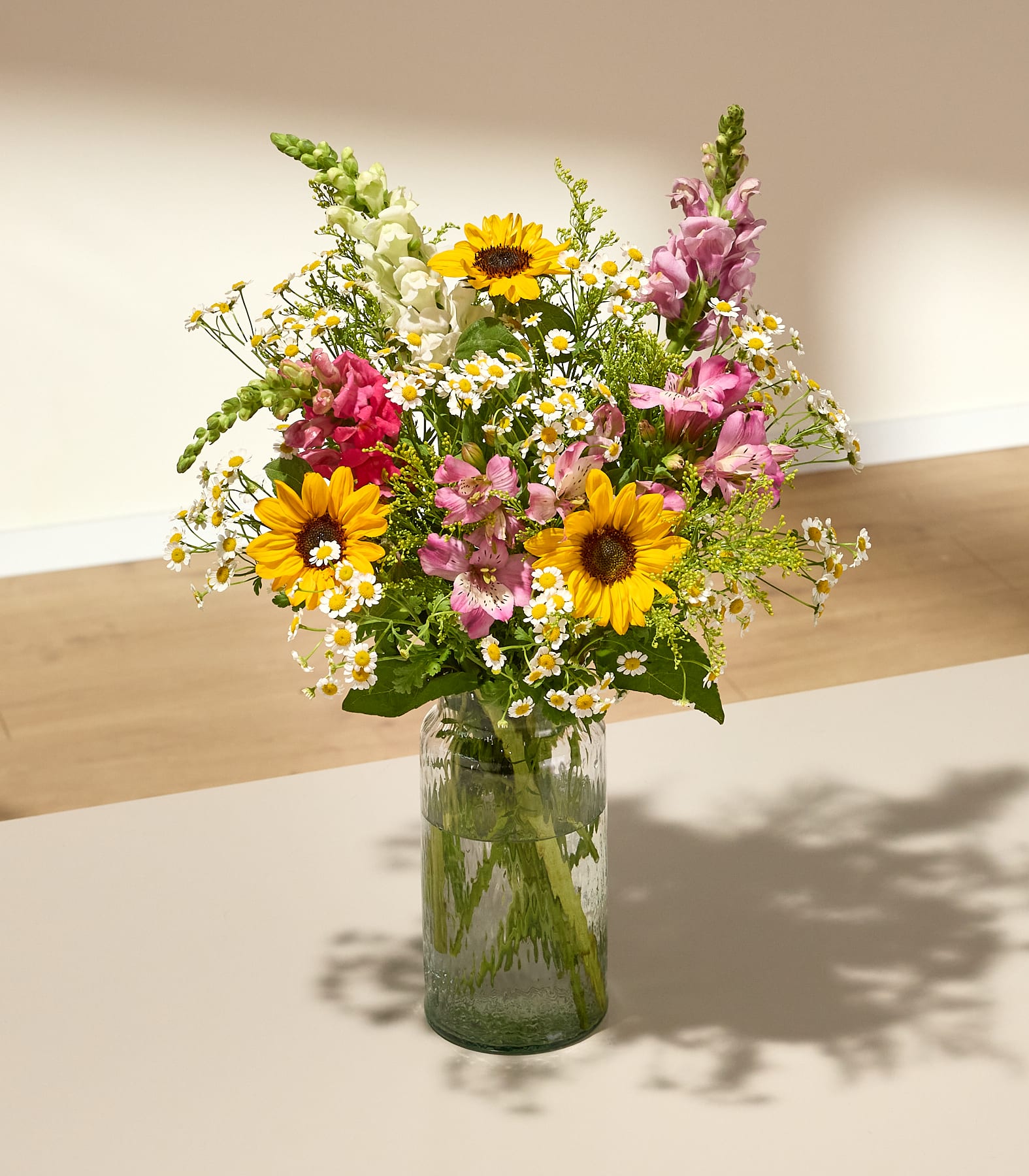 Ein farbenfroher Strauß aus Sonnenblumen, Löwenmäulchen und Gänseblümchen in einer Glasvase steht auf einer hellen Oberfläche; im Hintergrund sind eine sanft beleuchtete Wand und ein Holzboden zu sehen.