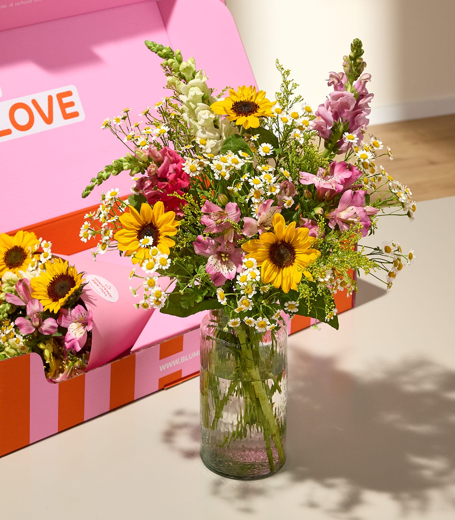 Ein farbenfroher Strauß aus Sonnenblumen, Löwenmäulchen und Gänseblümchen in einer Glasvase steht vor einer rosa Schachtel mit der Aufschrift „LOVE“.