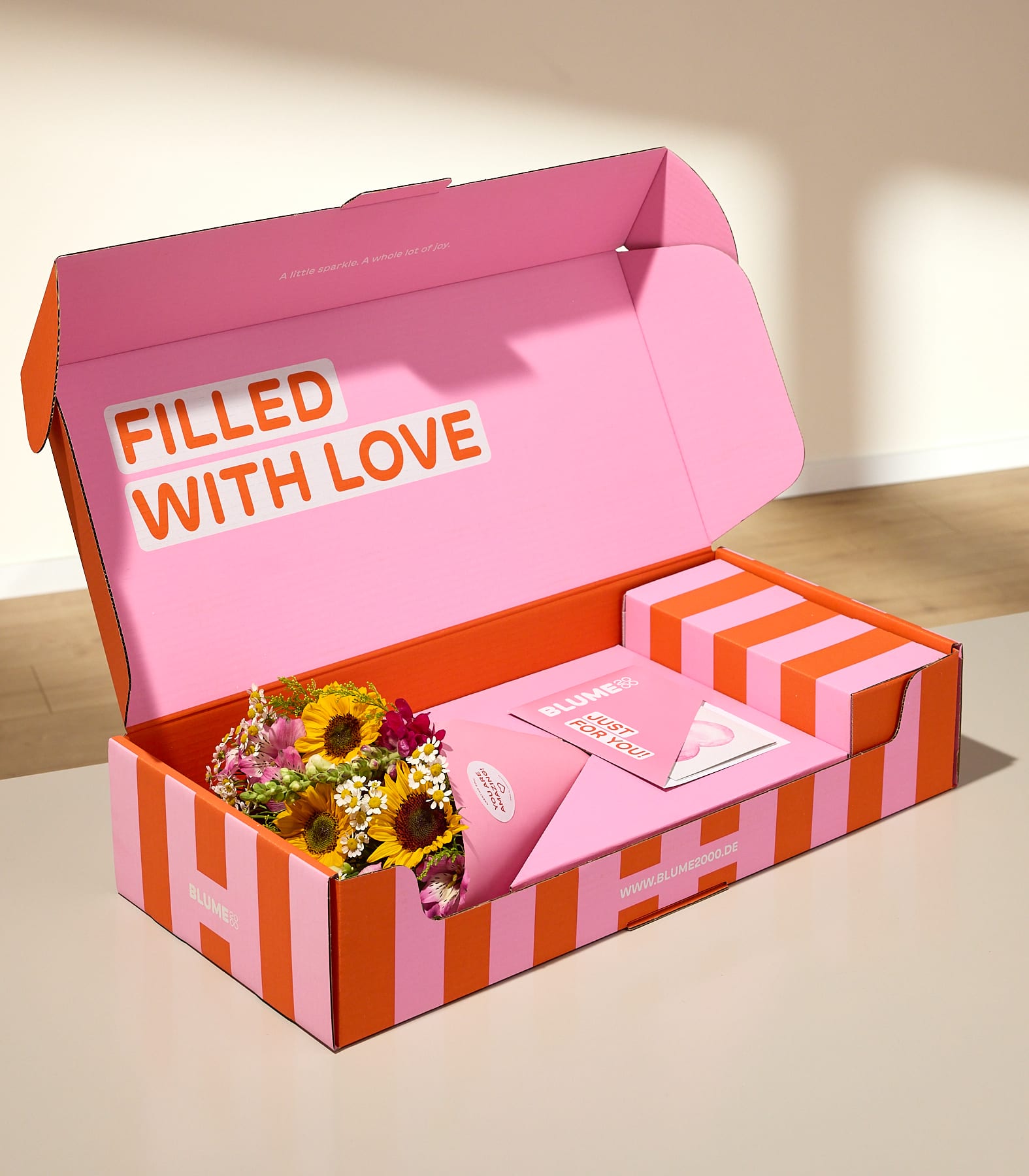 Eine rosa-orange gestreifte Schachtel mit der Aufschrift „FILLED WITH LOVE“ im Inneren enthält einen Sonnenblumenstrauß und eine Karte vor einem sanft beleuchteten Hintergrund.