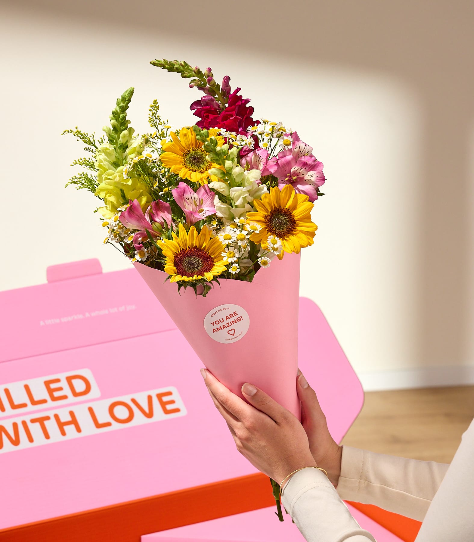 In Händen hält man einen leuchtenden Strauß Sonnenblumen und anderer bunter Blumen, eingewickelt in rosa Papier, neben einer hellrosa Schachtel mit der Aufschrift „VON LIEBE GEFÜLLT“.