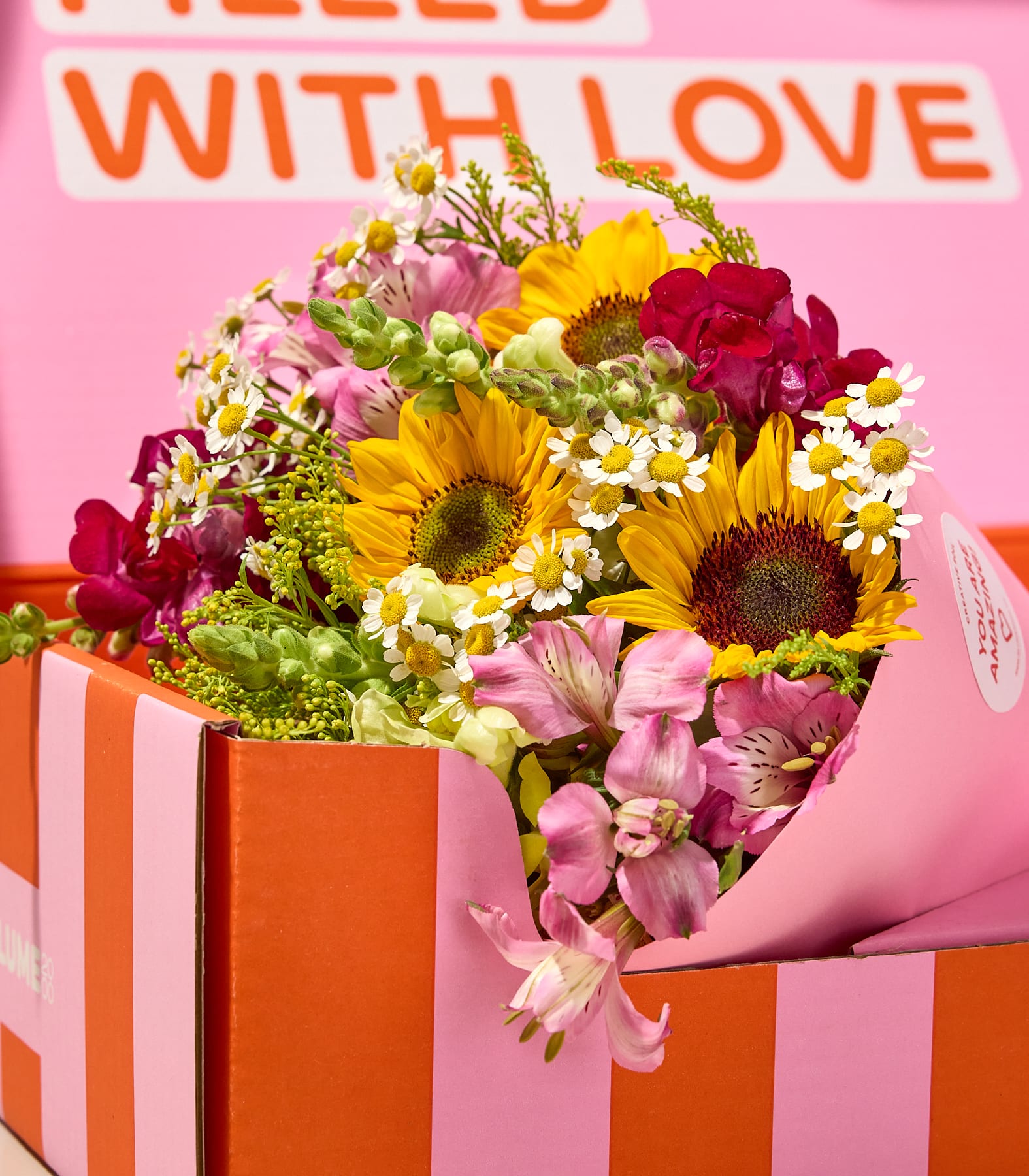 Aus einer gestreiften orange-rosa Schachtel lugt ein leuchtender Strauß aus Sonnenblumen, Gänseblümchen und Alstroemerien hervor; im rosa Hintergrund sind die Worte „FILLED WITH LOVE“ zu erkennen.