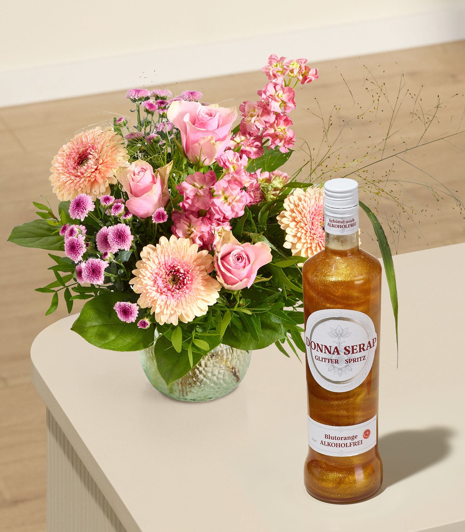 Ein leuchtender Strauß rosa Blumen steht in einer Glasvase neben einer Flasche prickelndem Goldlikör auf einer hellen Oberfläche, im Hintergrund sind Wand und Boden verschwommen zu sehen.