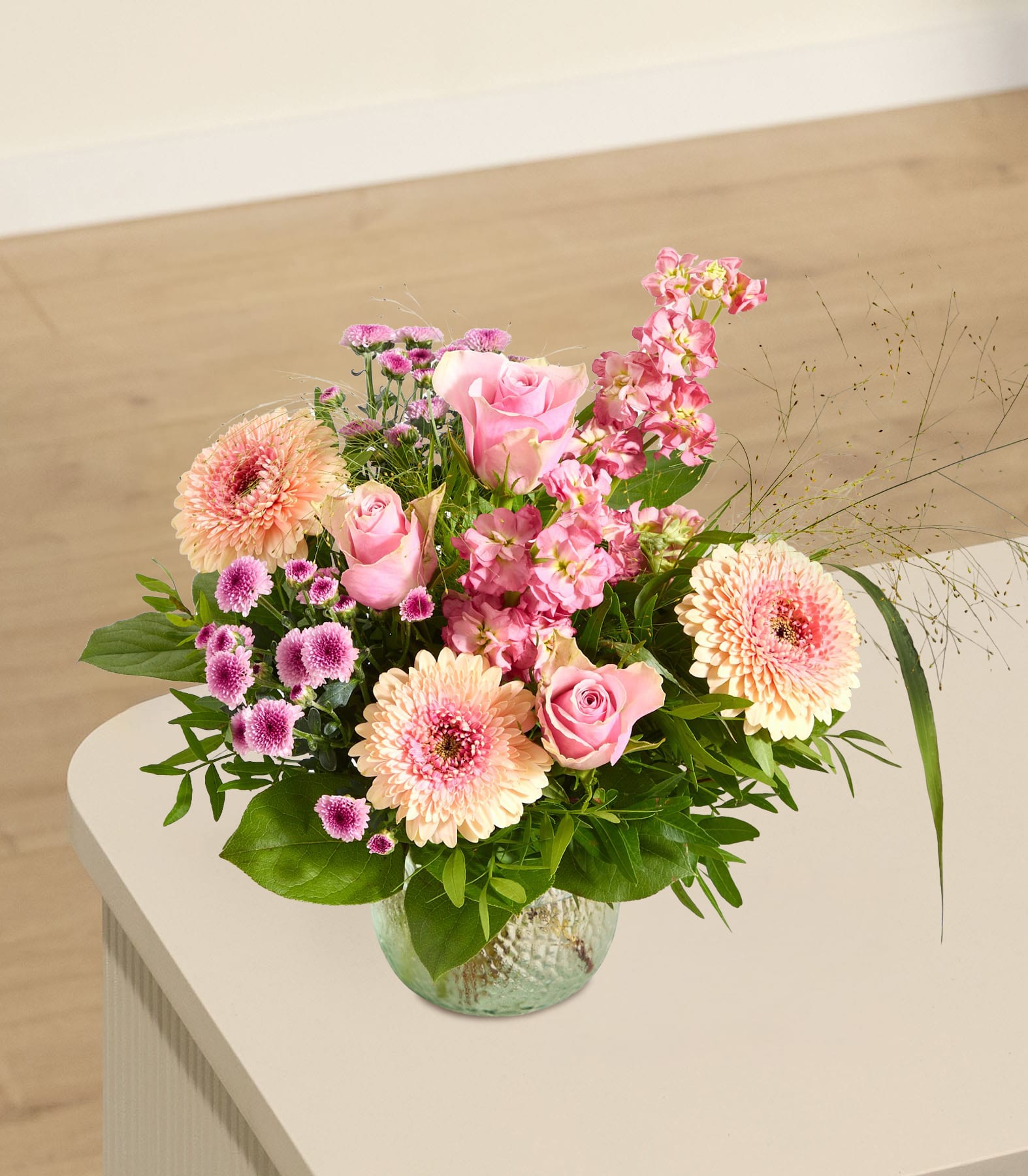 Ein leuchtender Strauß rosa und pfirsichfarbener Blumen steht in einer strukturierten Glasvase auf einer hellen Oberfläche, im Hintergrund sind Wand und Boden sanft verschwommen zu sehen.