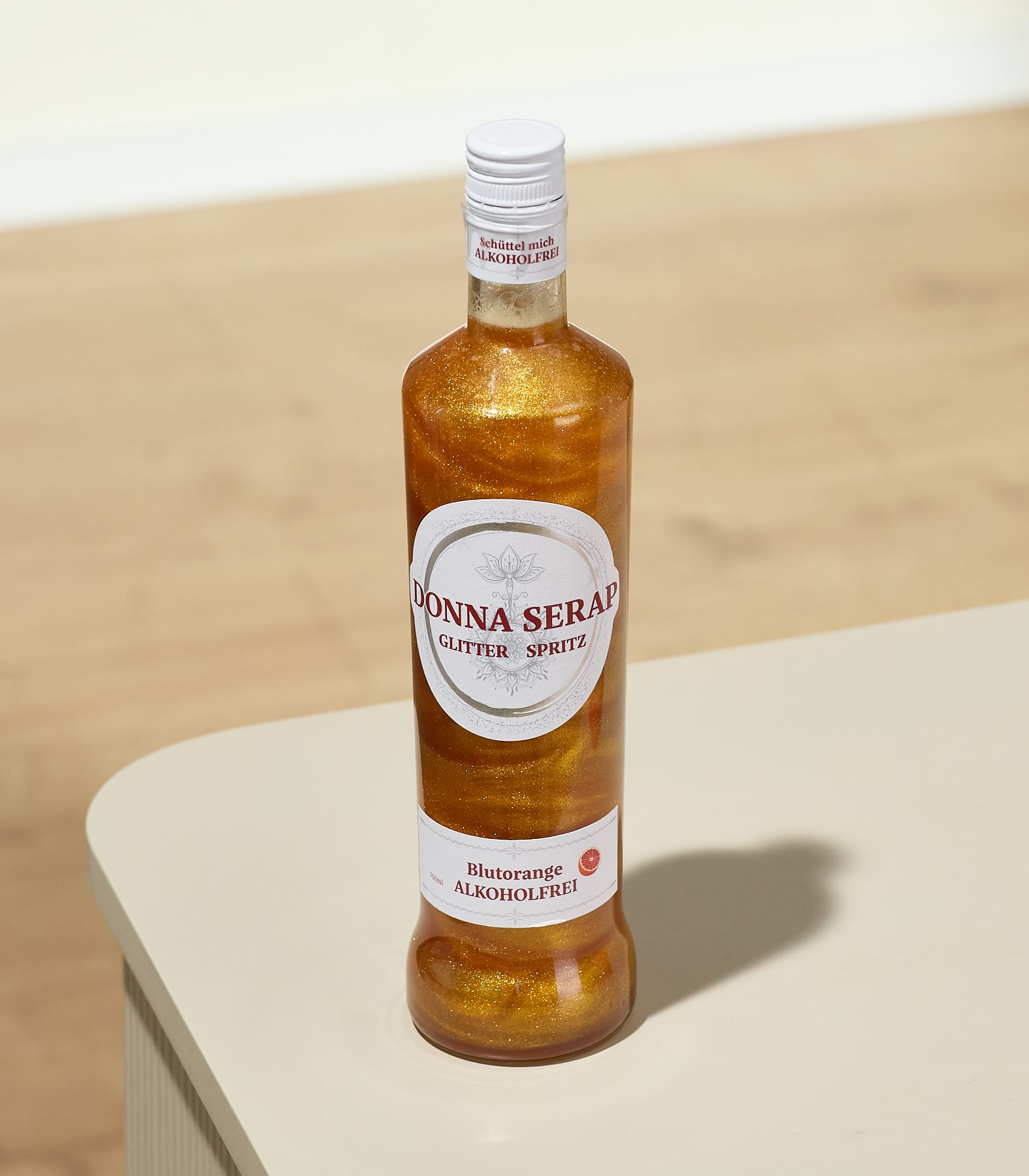 Eine Flasche glitzernder Orangen-Spritzer steht auf einem hellen Tisch vor einem verschwommenen Hintergrund aus Holzboden und -wand.