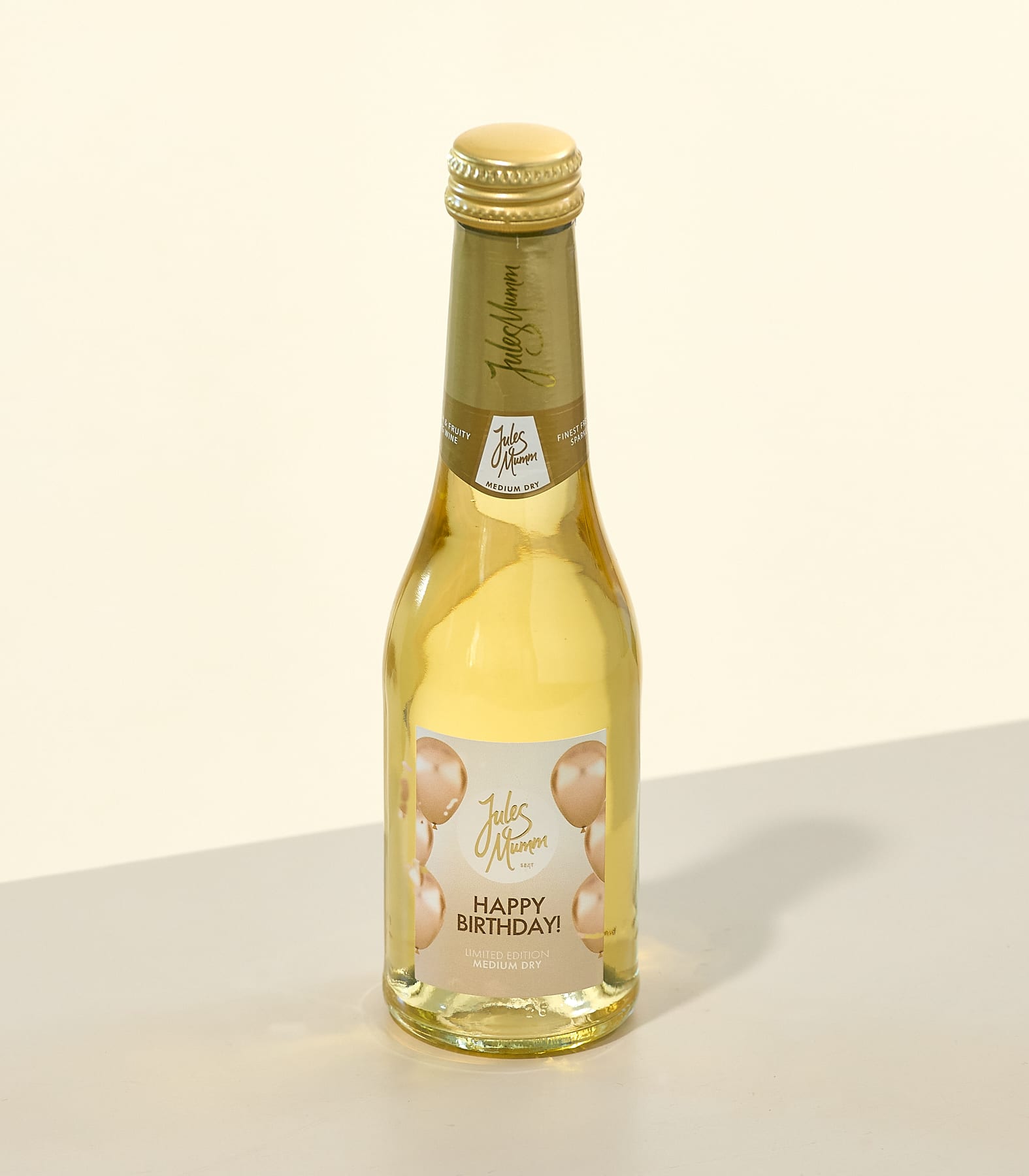Eine goldfarbene Glasflasche mit der Aufschrift „Happy Birthday“ steht auf einer hellen Oberfläche vor einem schlichten weißen Hintergrund.