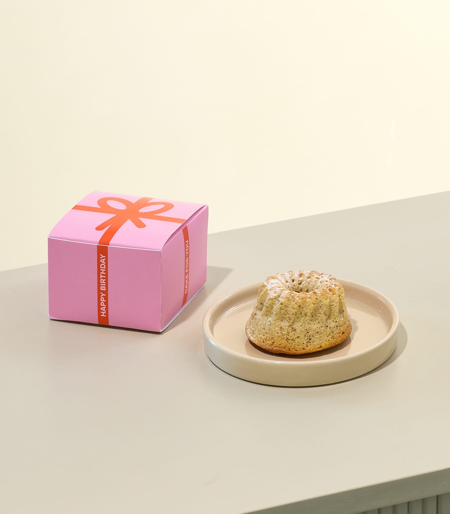 Auf einer Holzoberfläche steht ein hellfarbiger Kuchen oder ein Gebäck, daneben eine hellblaue quadratische Schachtel mit der Aufschrift „HAPPY BIRTHDAY“.
