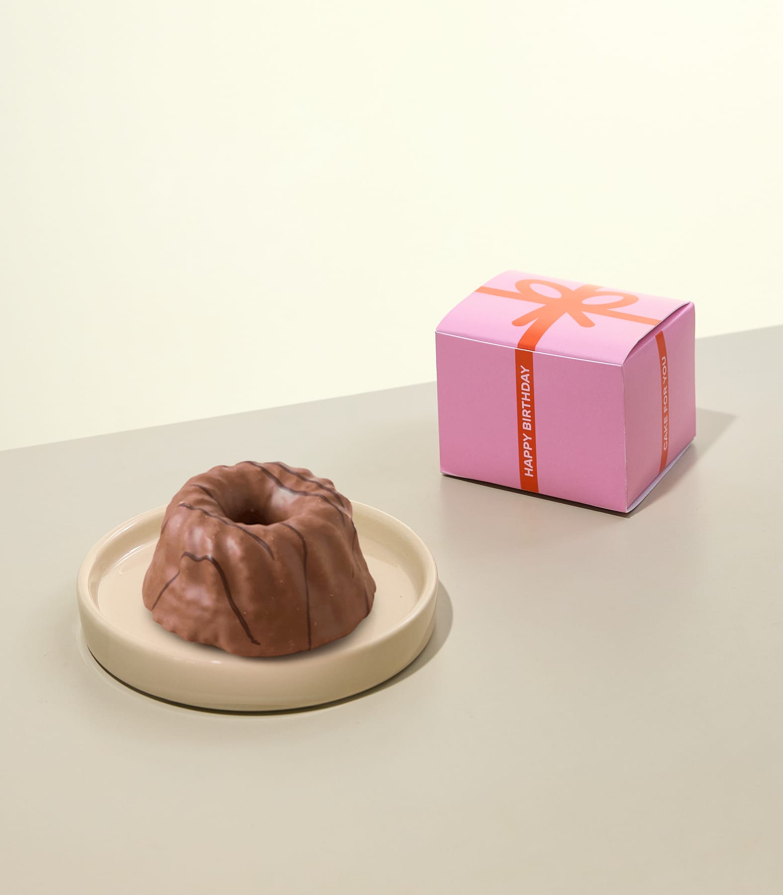 Auf einem hellrosa Teller steht ein kleiner, runder Schokoladenkuchen; im Hintergrund ist eine grün-weiße Schachtel mit der Aufschrift „Kuchen für dich“ zu sehen. (Beschreibungstext KI-generiert)