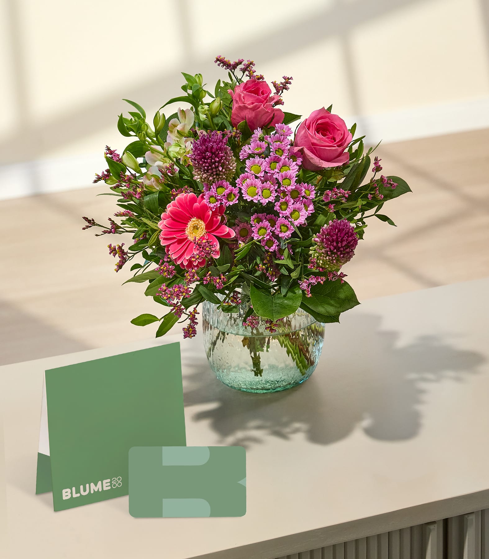 Ein leuchtender Strauß rosa Blumen in einer Glasvase steht auf einer hellen Oberfläche vor einem sanft beleuchteten Hintergrund mit Schatten. (Beschreibungstext KI-generiert)