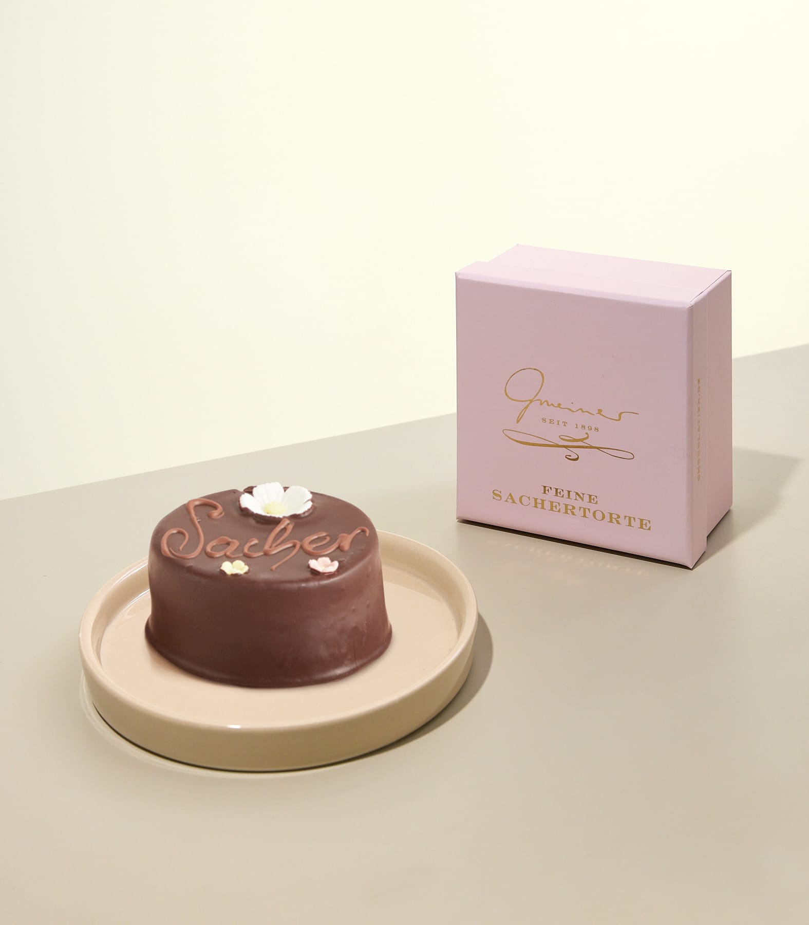 Auf einem hellen Teller steht ein kleines, rundes Schokoladendessert, daneben eine Schachtel mit der Aufschrift „Gourmet“ vor einem schlichten, hellen Hintergrund.