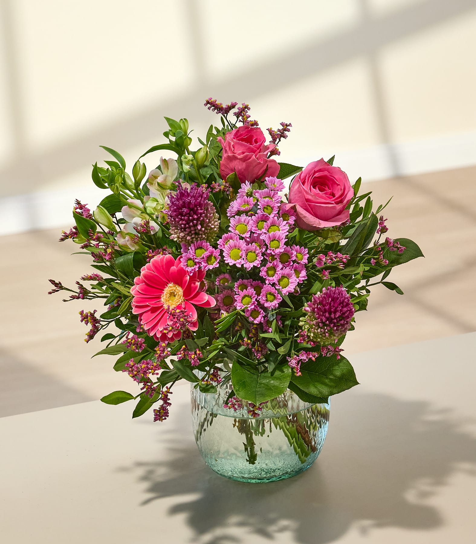 Ein leuchtender Strauß rosa und violetter Blumen in einer durchsichtigen Glasvase steht auf einer hellen Oberfläche, während sanftes Sonnenlicht Schatten auf eine helle Wand im Hintergrund wirft. (Beschreibungstext KI-generiert)