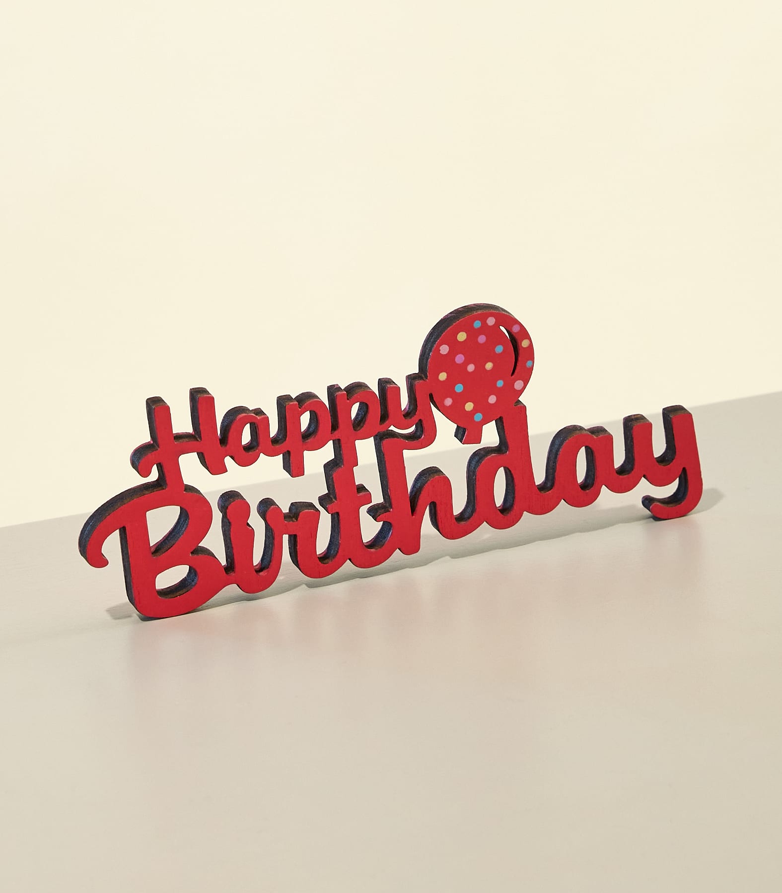 Ein roter „Happy Birthday“-Kuchenaufsatz mit einem gepunkteten Ballon steht auf einer hellen Oberfläche vor einem sanften, cremeweißen Hintergrund. (Beschreibungstext KI-generiert)