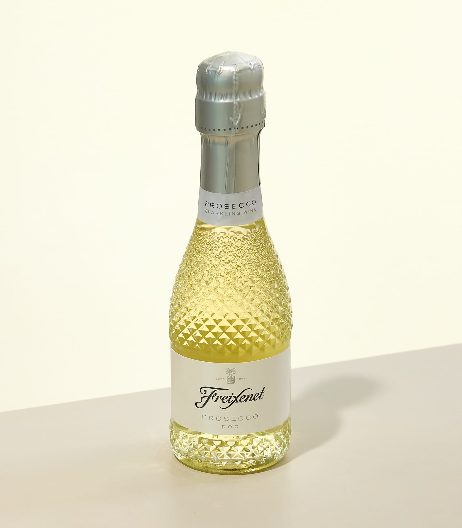 Eine kleine Flasche Freixenet Prosecco mit strukturiertem Glaskörper steht auf einer hellen Oberfläche vor einem blassgelben Hintergrund. (Beschreibungstext KI-generiert)