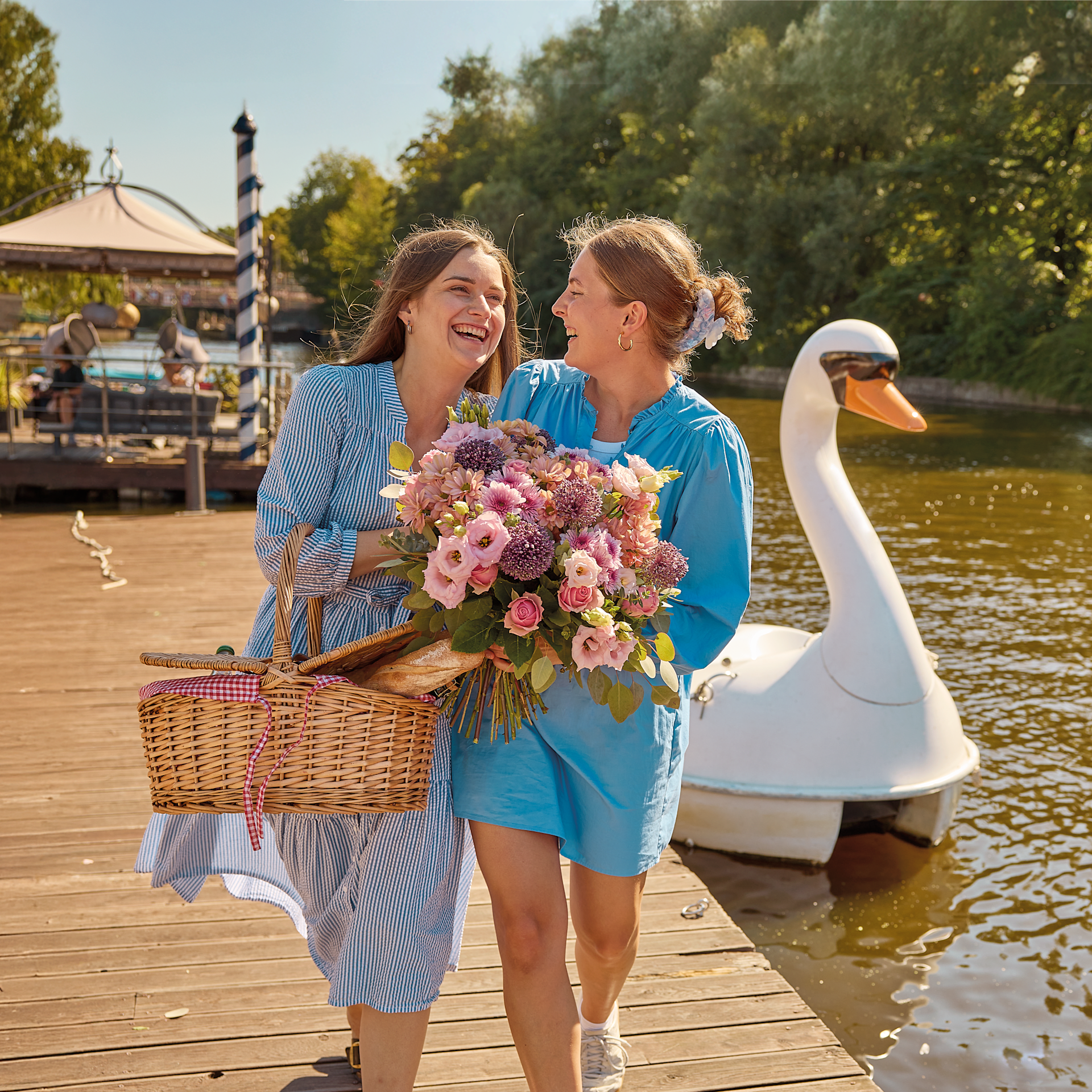 Zwei Frauen mit einem Picknickkorb und Blumen spazieren auf einem Holzsteg, im Wasser befindet sich ein Schwanenboot, im Hintergrund sind Bäume zu sehen. (Beschreibungstext KI-generiert)