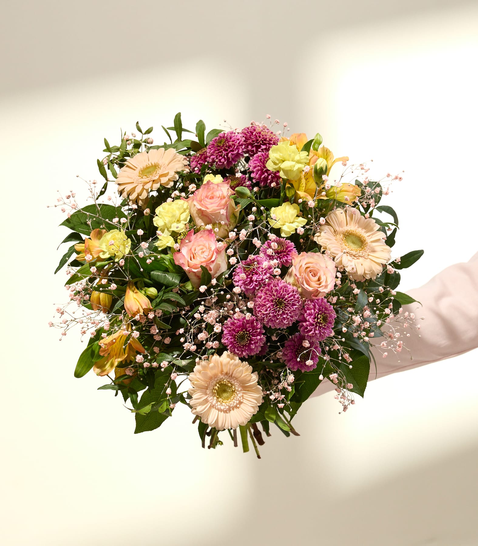 Ein leuchtender Strauß aus rosa, gelben und pfirsichfarbenen Blüten mit zartem Schleierkraut hebt sich vor einem sanft beleuchteten, weißen Hintergrund mit einem dezenten Schatten ab.