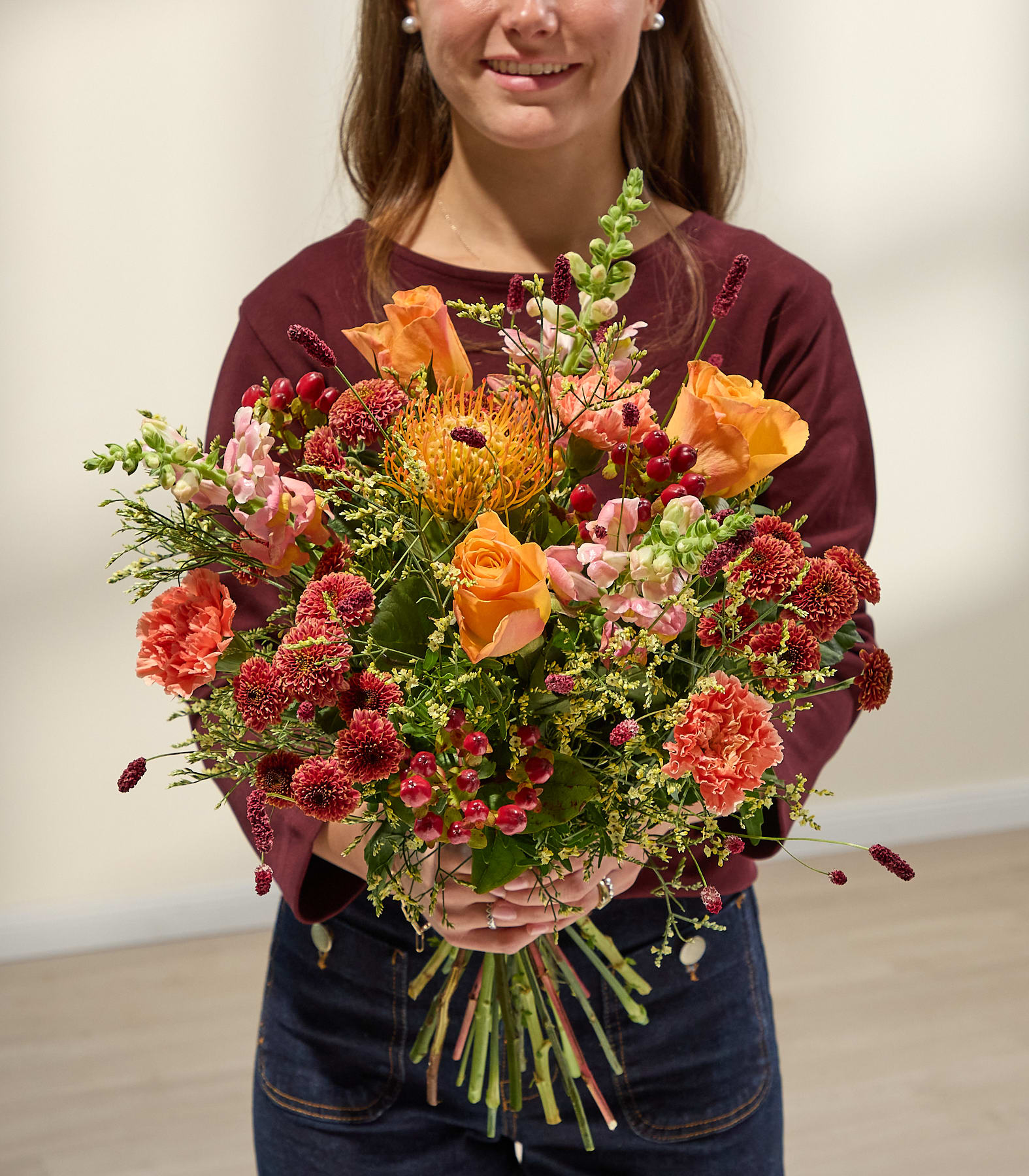 Eine Person in einem weinroten Hemd und Jeans hält einen leuchtenden Strauß orangefarbener und rosafarbener Blumen vor einem schlichten, hellen Hintergrund. (Beschreibungstext KI-generiert)