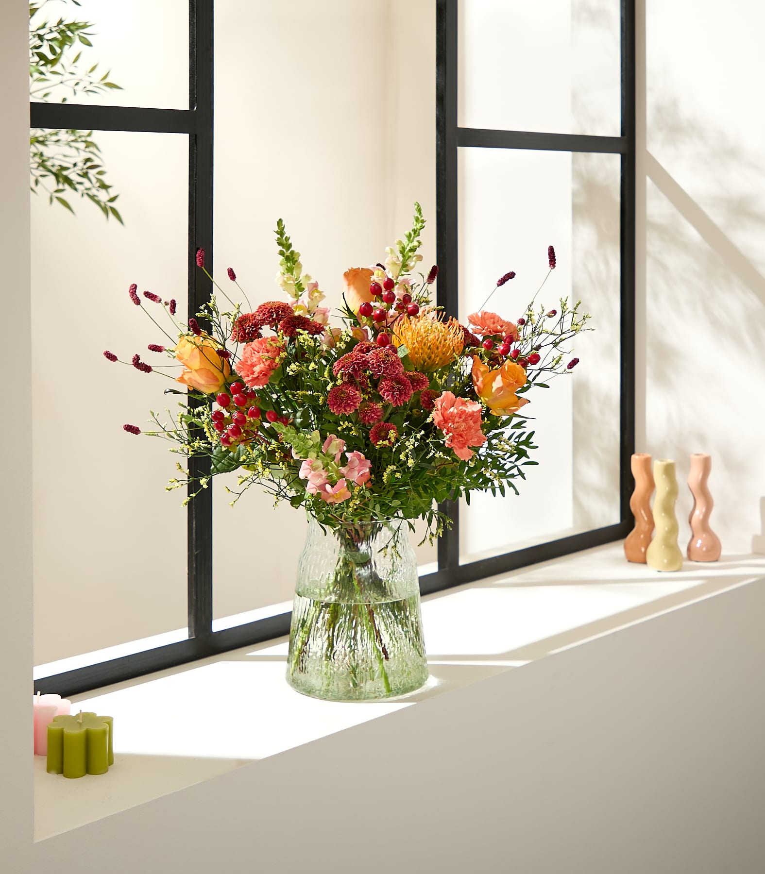 Auf einem Fensterbrett steht ein farbenfroher Strauß herbstlicher Blumen in einer strukturierten Glasvase, im Vordergrund dekorative Kerzen und im Hintergrund ein Fenster mit gefiltertem Licht. (Beschreibungstext KI-generiert)