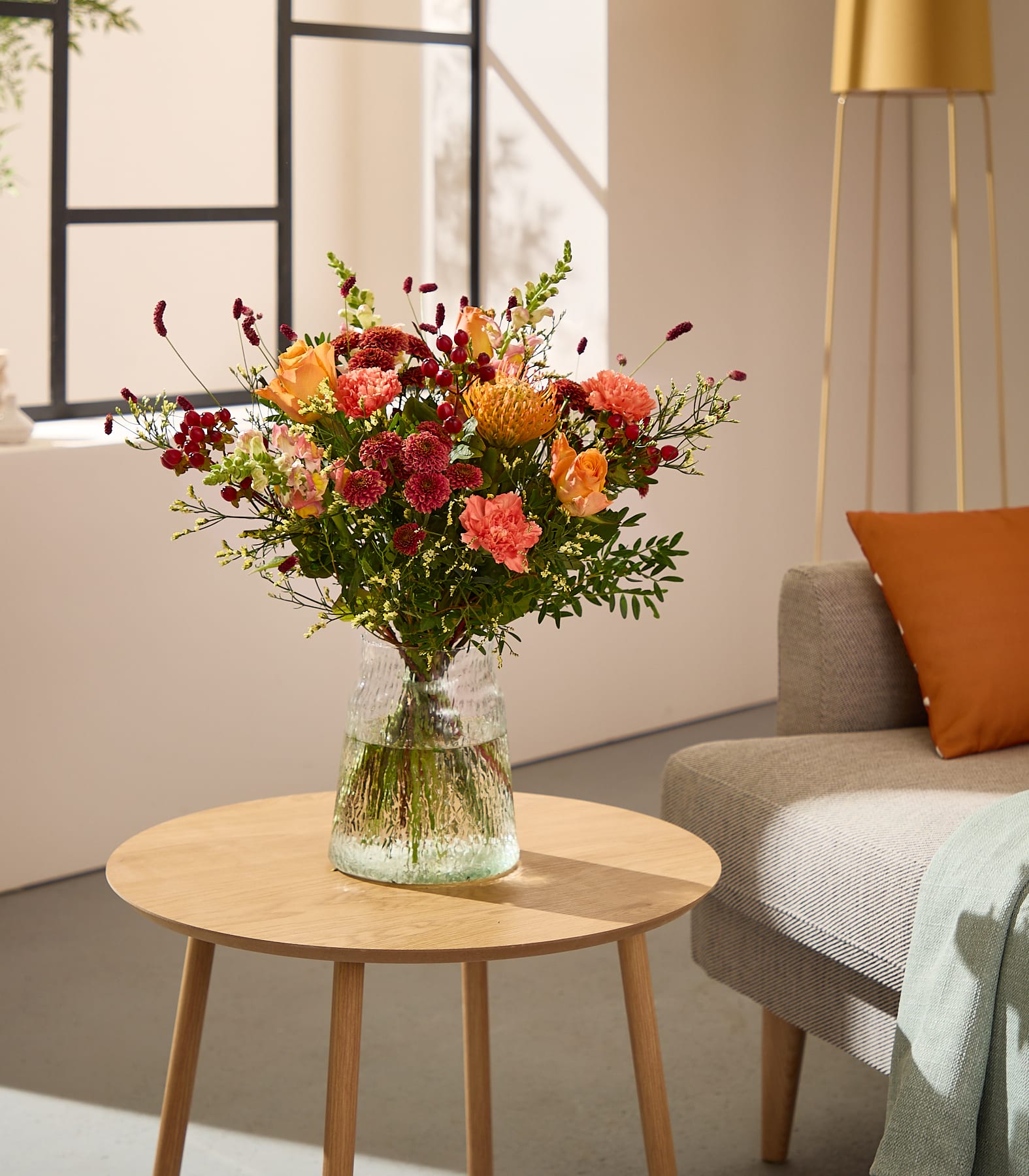 Im Vordergrund steht ein leuchtender Strauß orangefarbener und roter Blumen in einer Glasvase auf einem Holztisch; im sanft beleuchteten Hintergrund sind ein Sofa und eine Stehlampe zu sehen. (Beschreibungstext KI-generiert)