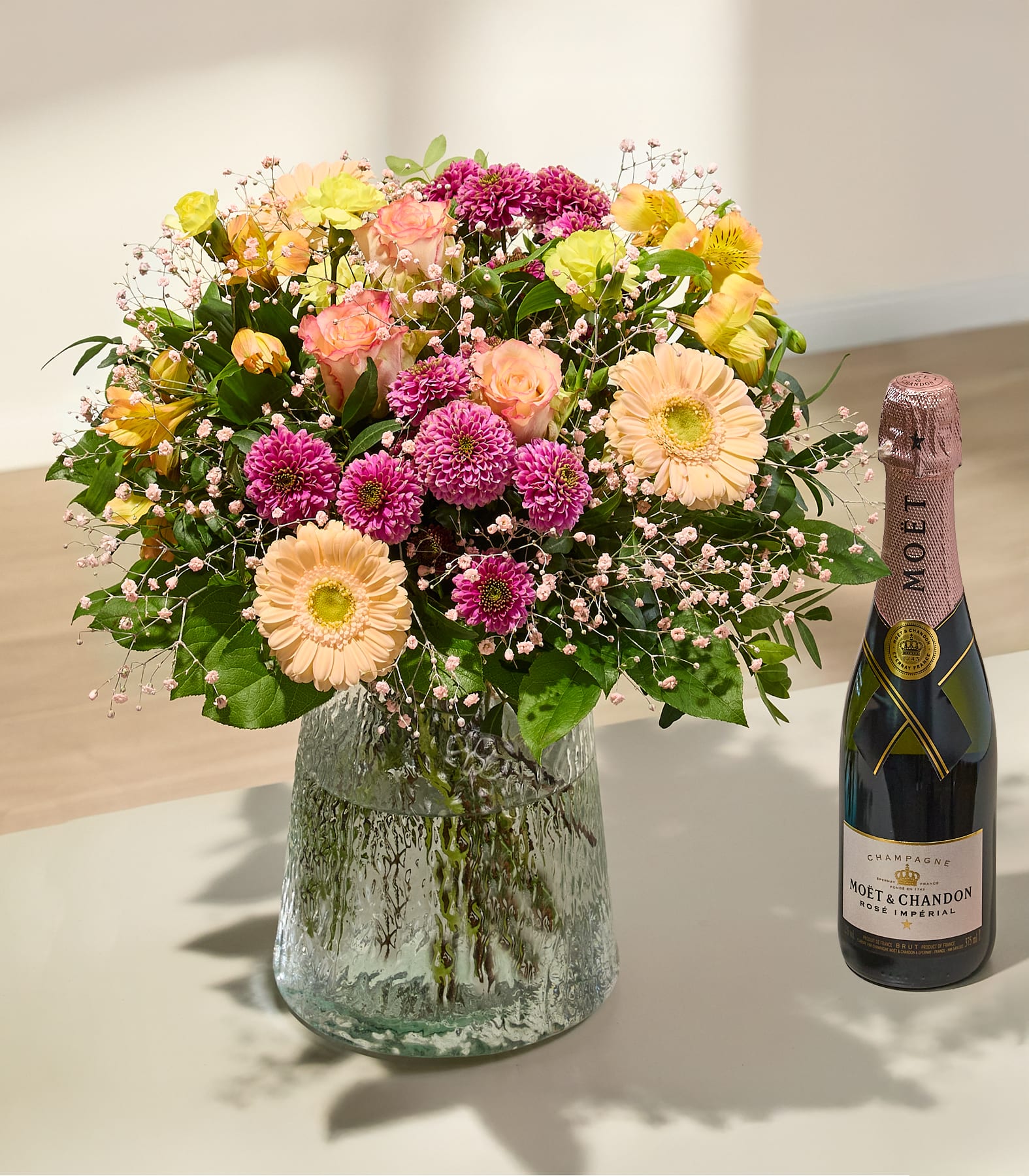 Ein farbenfroher Strauß aus rosa, gelben und weißen Blumen steht neben einer Flasche Sekt vor einem schlichten weißen Hintergrund.