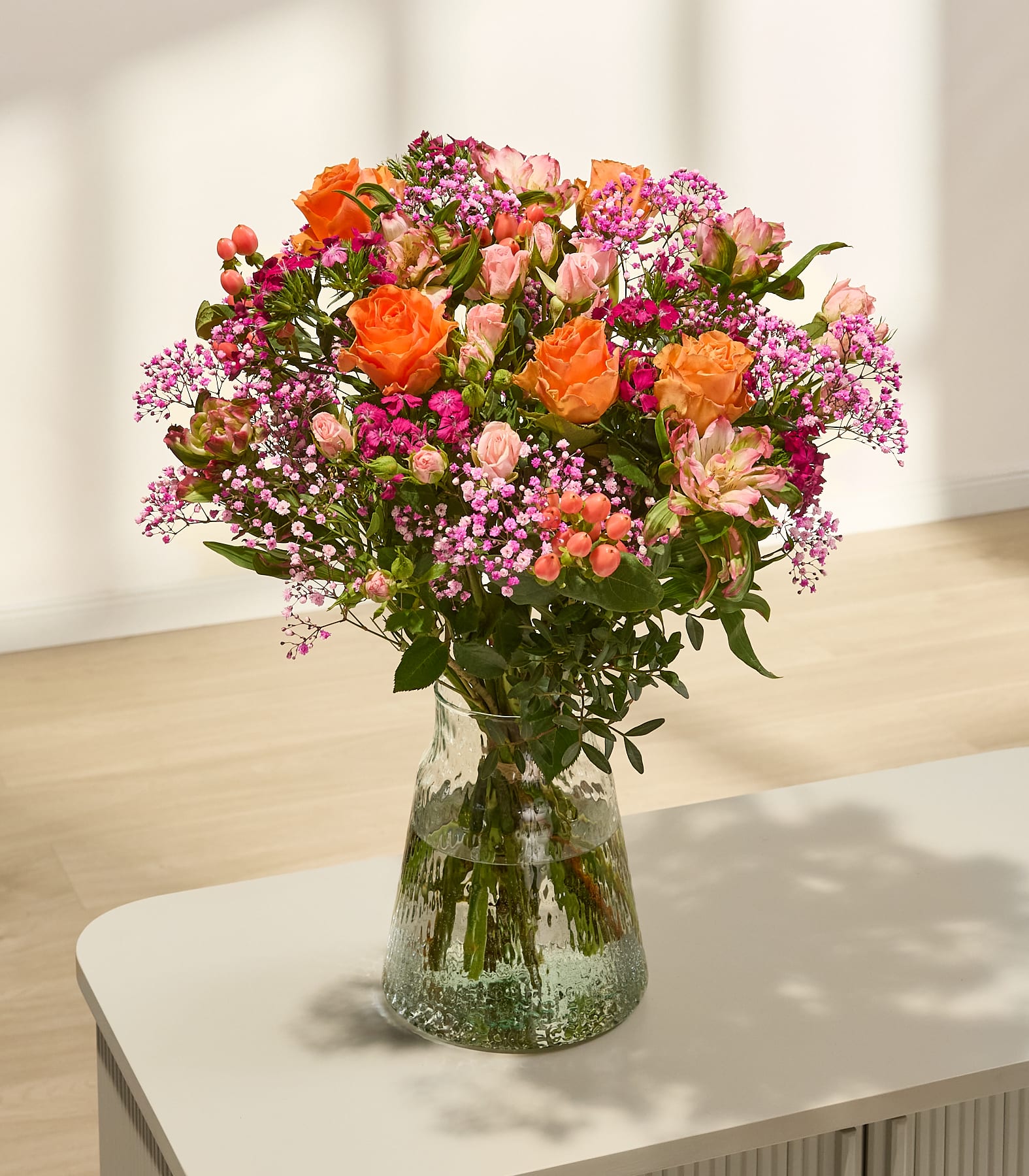 Ein lebhafter Strauß bunter Blumen, darunter rosa und gelbe Rosen, weiße Gänseblümchen und andere Blüten, in den Händen einer Person vor einem schlichten, hellen Hintergrund. (Beschreibungstext KI-generiert)