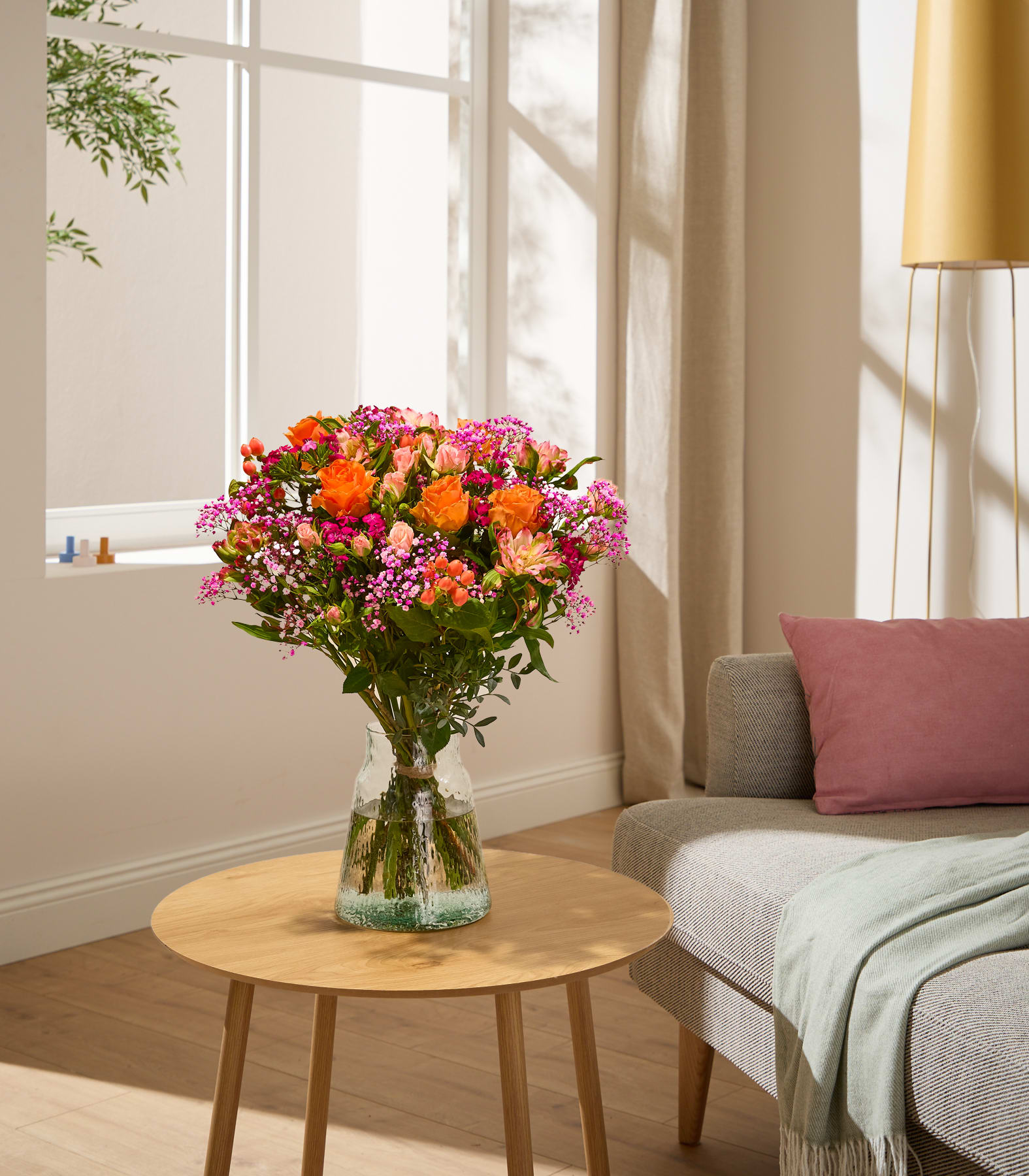 Im Vordergrund steht ein leuchtender Strauß orangefarbener und rosafarbener Blumen in einer Glasvase auf einem Holztisch; im Hintergrund sind ein sonnendurchfluteter Raum und eine Couch zu sehen. (Beschreibungstext KI-generiert)