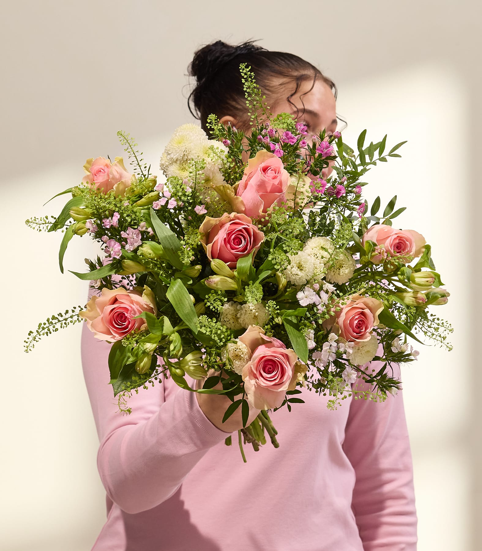 Eine Person in einem rosa Pullover hält vor einem schlichten Hintergrund einen farbenfrohen Strauß aus rosa Rosen, weißen Blüten und Grünzeug. (Beschreibungstext KI-generiert)