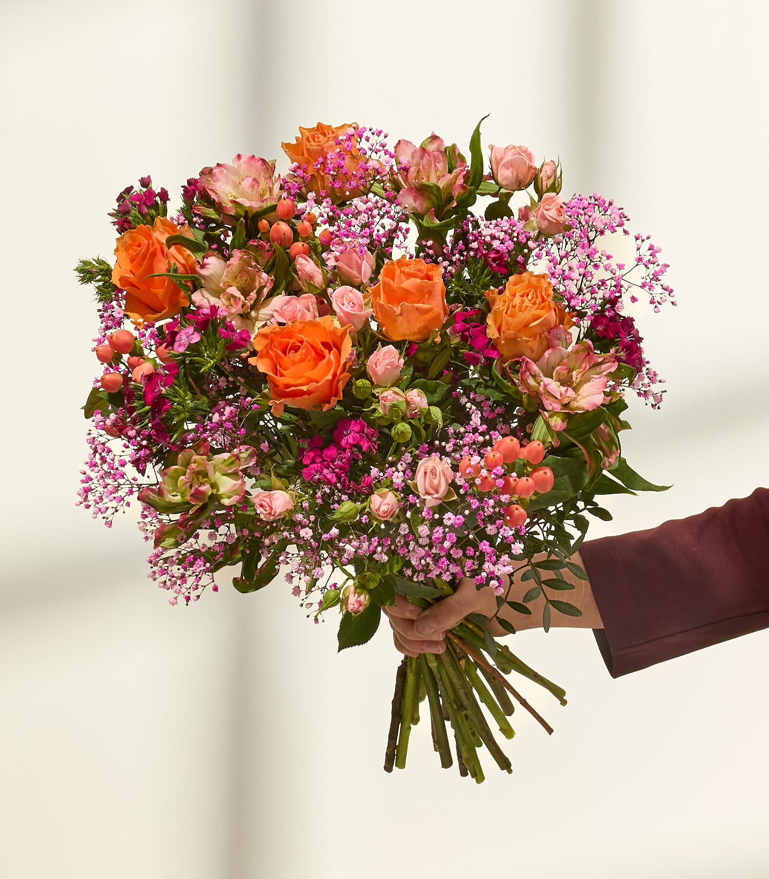 Ein farbenfroher Blumenstrauß, darunter orangefarbene Rosen, rosa Nelken und lila Strandflieder, in den Händen einer Person vor einem schlichten Hintergrund. (Beschreibungstext KI-generiert)