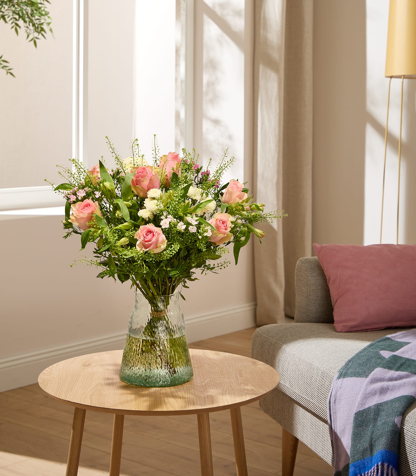 Eine Frau in einem rosa Pullover hält einen großen Strauß bunter Blumen, darunter rosa Rosen, weiße Chrysanthemen und anderes Grün, vor einem schlichten weißen Hintergrund. (Beschreibungstext KI-generiert)