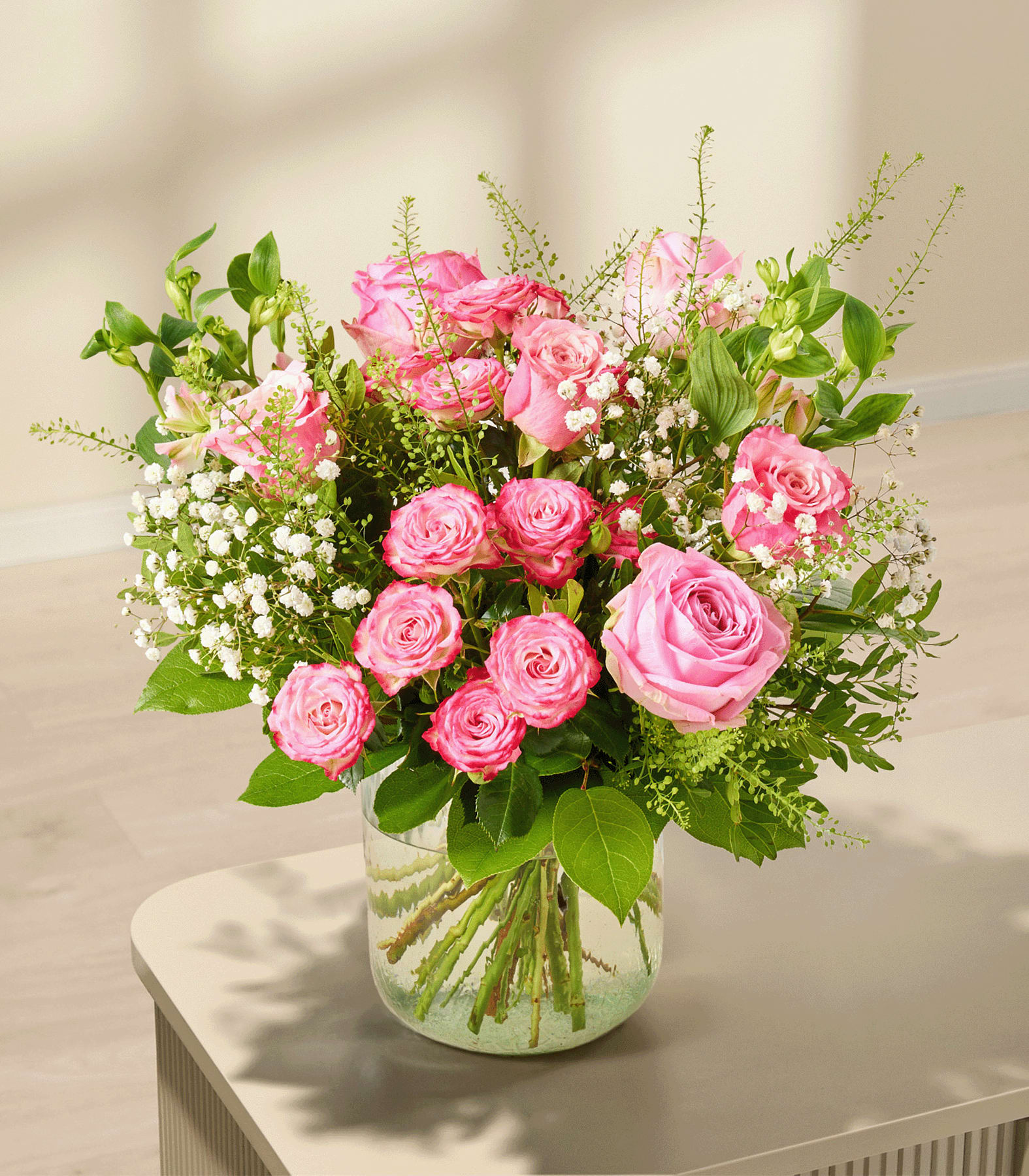 Ein Strauß rosa Rosen und Schleierkraut steht in einer Glasvase auf einem hellen Tisch, im Hintergrund ist eine sanft beleuchtete Wand zu sehen.