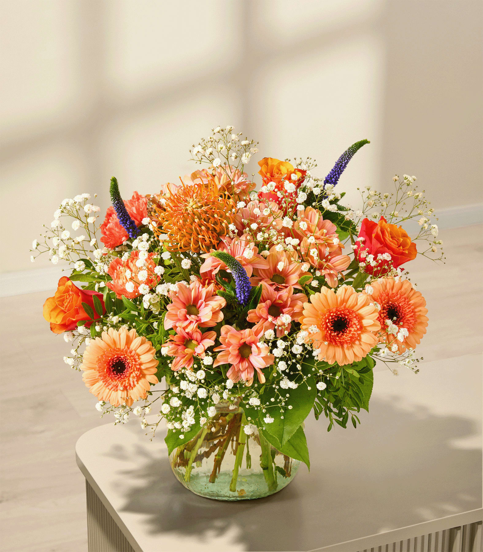Ein leuchtender Strauß aus orangefarbenen und rosafarbenen Blumen mit zartem weißen Schleierkraut füllt eine Glasvase auf einer hellen Oberfläche vor einem sanft beleuchteten Hintergrund mit gefleckten Schatten. (Beschreibungstext KI-generiert)