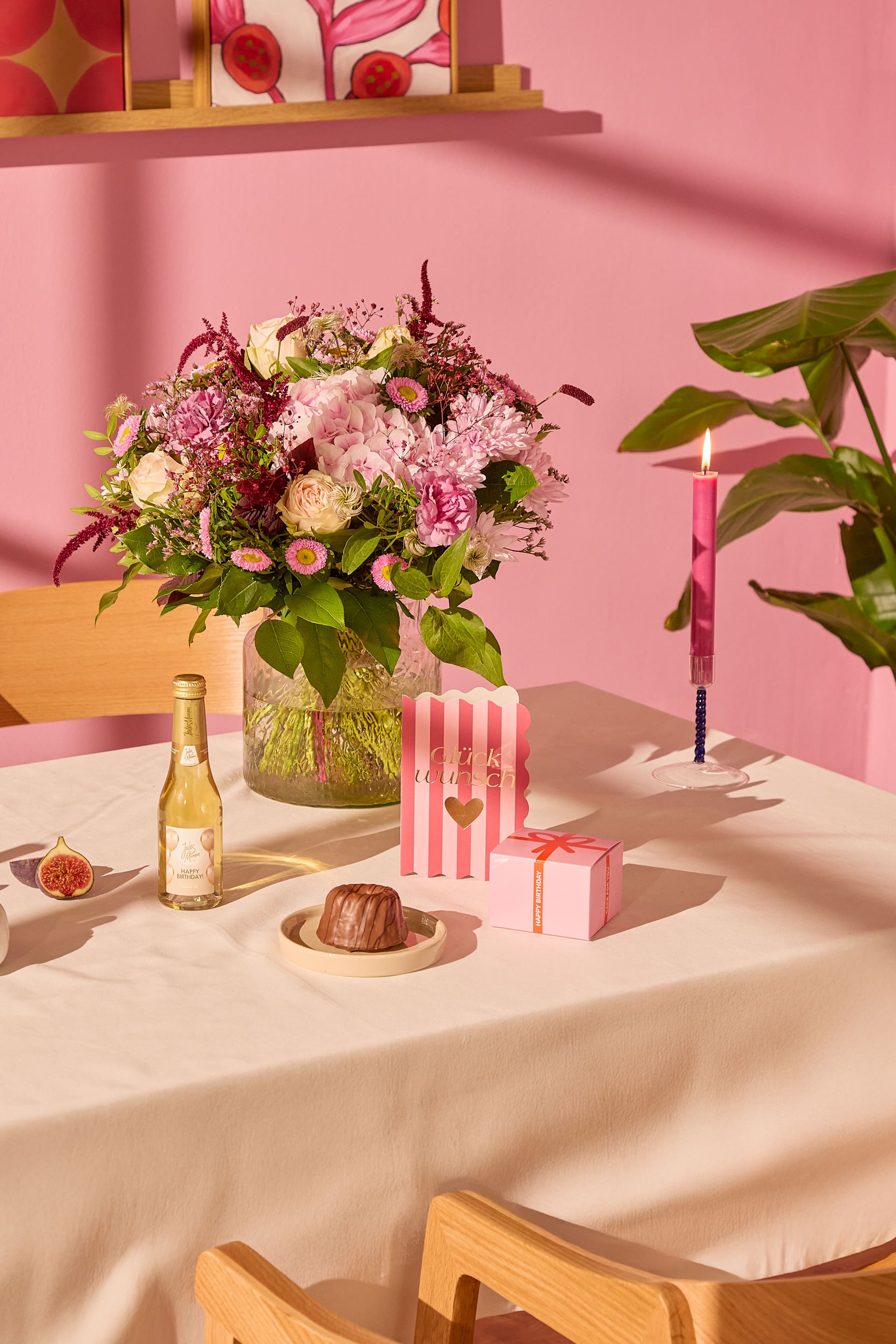 Ein mit Blumenarrangements, Geschenken und einer Kerze gedeckter Tisch liegt vor einer rosa Wand in sanftem rosa Licht. (Beschreibungstext KI-generiert)