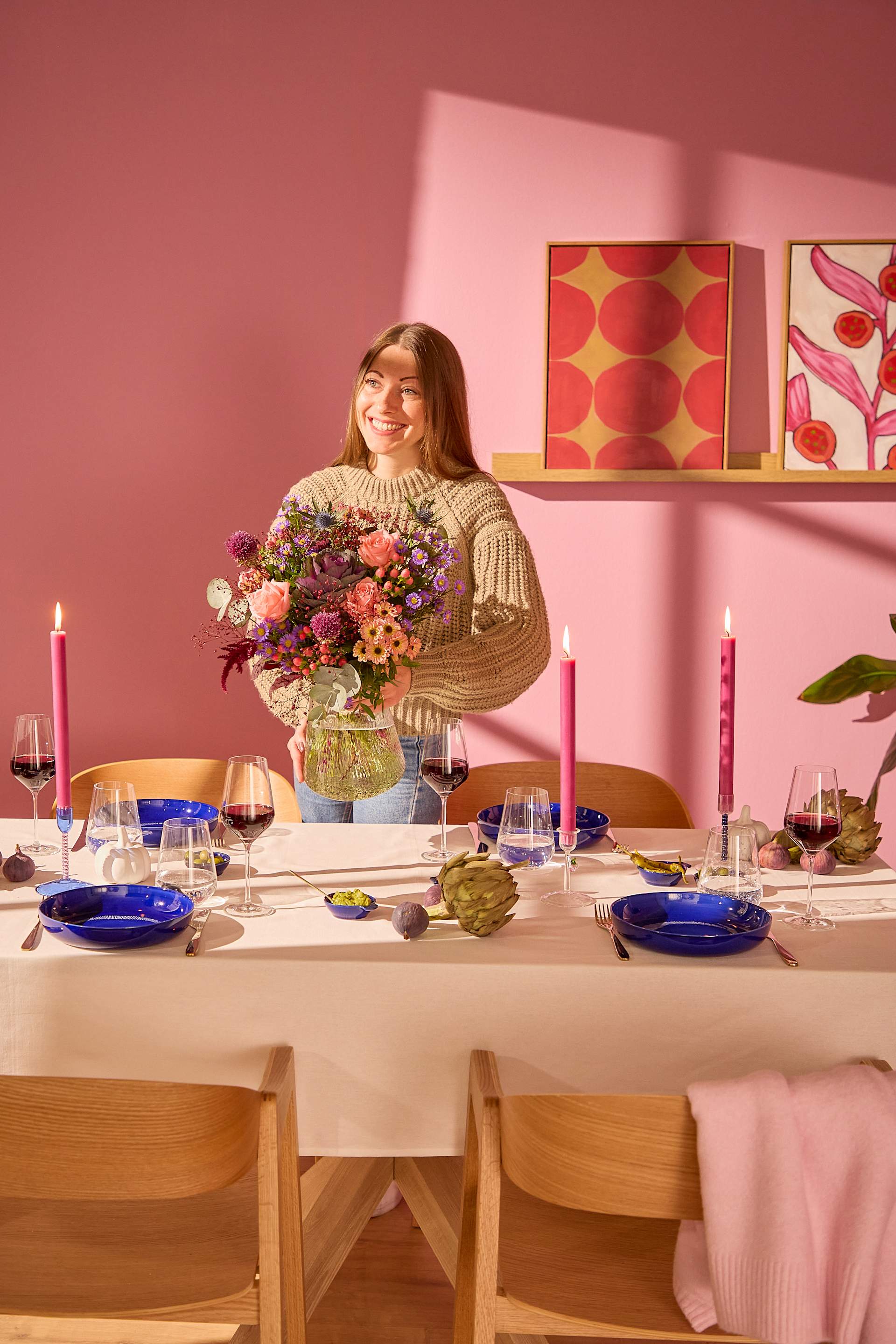 Eine Frau hält einen Blumenstrauß über einen zum Abendessen gedeckten Tisch mit rosa Kerzen und blauen Tellern vor einer rosa Wand mit gerahmten Bildern. (Beschreibungstext KI-generiert)