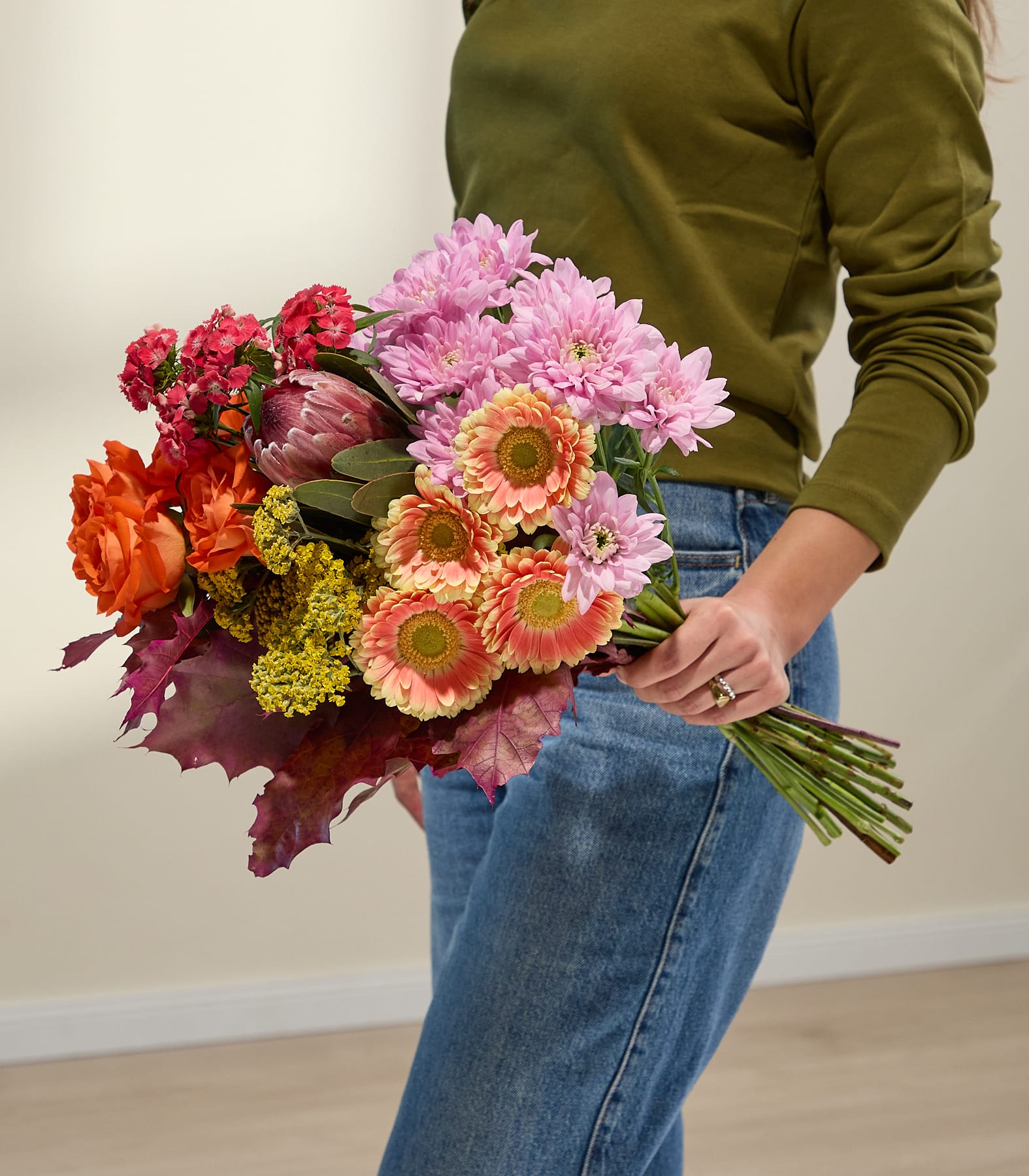 Eine Hand in einem grünen Ärmel hält einen leuchtenden Strauß aus orangefarbenen Rosen, rosa Chrysanthemen und anderen farbenfrohen Blumen vor einem sanft beleuchteten, neutralen Hintergrund. (Beschreibungstext KI-generiert)